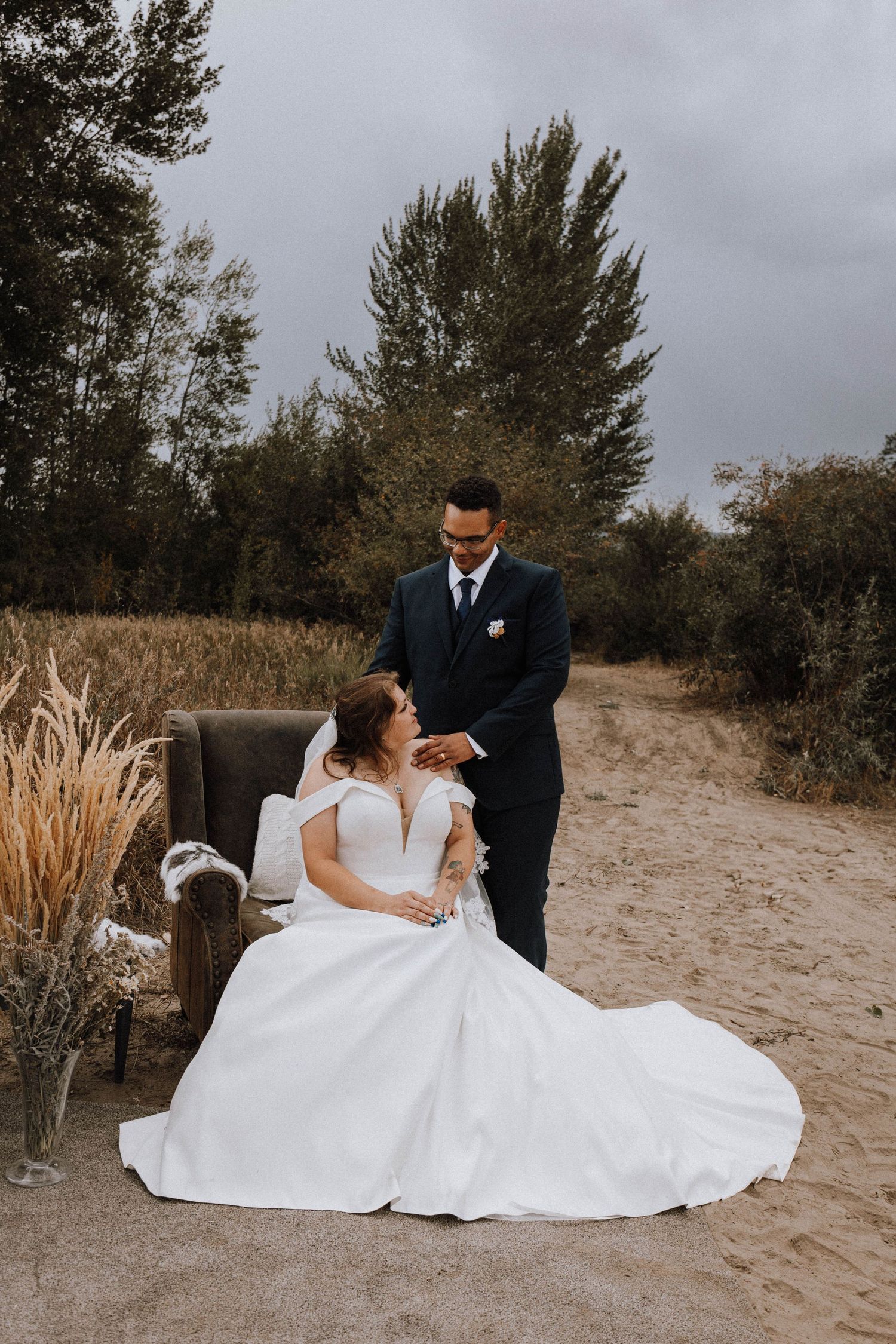 A romantic wedding photo on a dirt path with pampas grass decor and evergreen trees in the background on an overcast day.