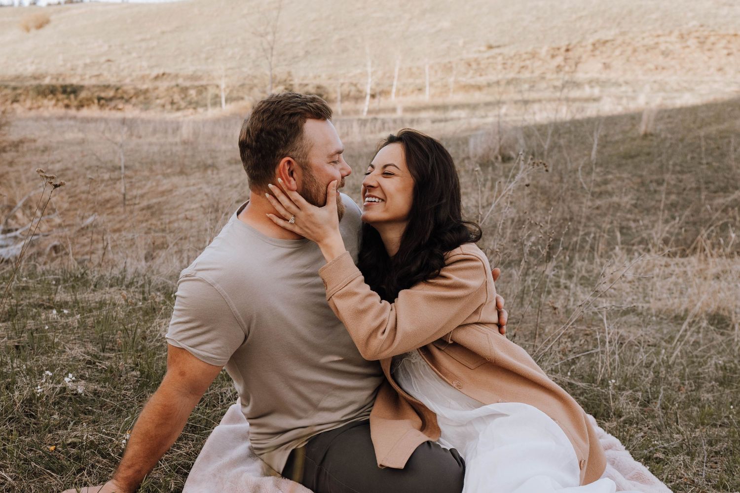 Two people sit together laughing intimately in a field at sunset with mountain backdrop.