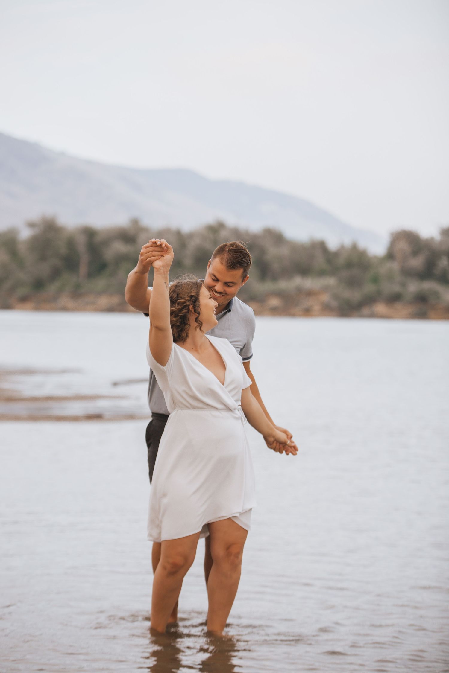 A couple dances together in shallow water with mountains and trees in the background.
