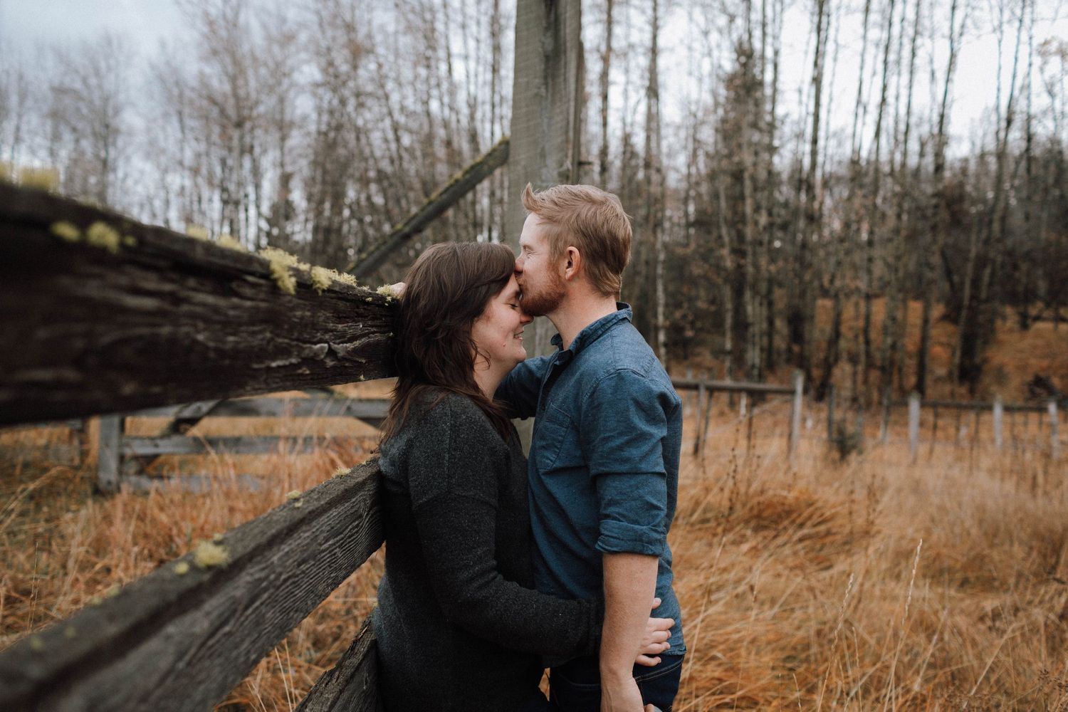 Intimate moment between couple against backdrop of fall trees and wooden fence.