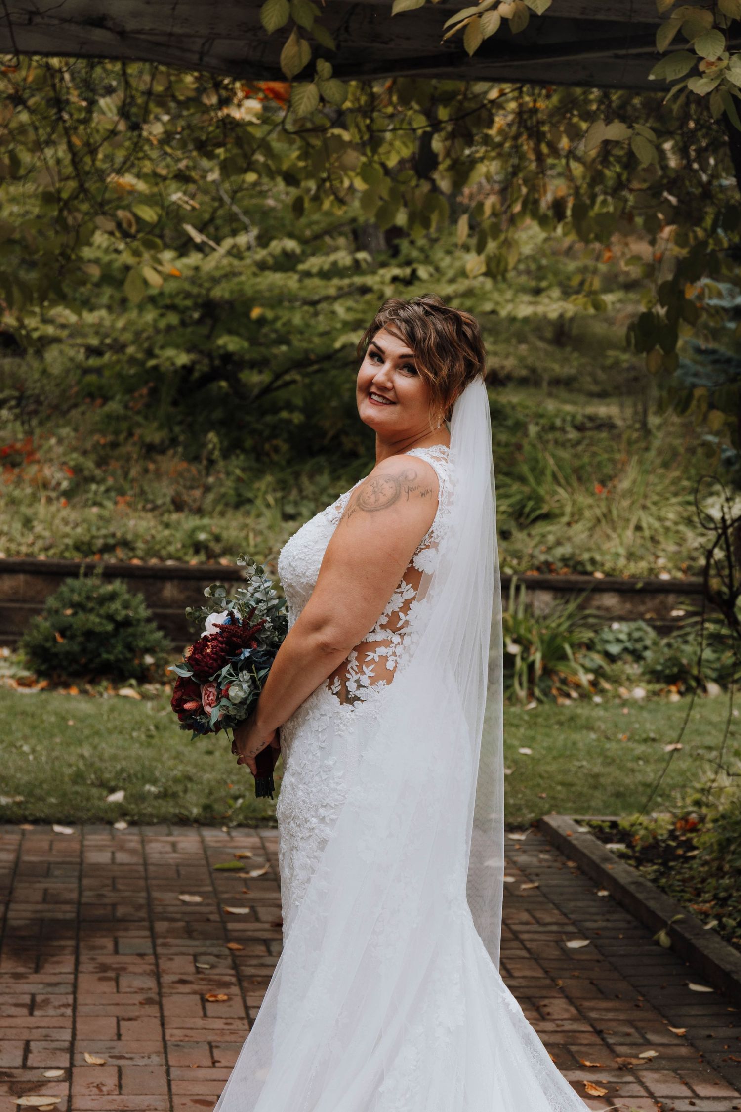 Bride in fitted lace gown poses with bouquet on brick pathway.