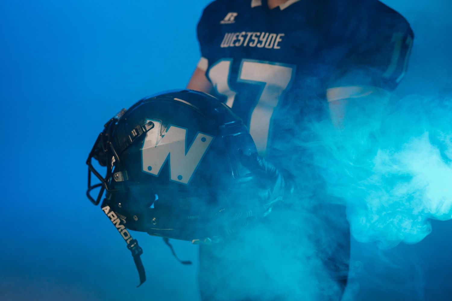 Football player in Westsyde jersey number 17 holds helmet amid blue smoke on dramatic dark background.