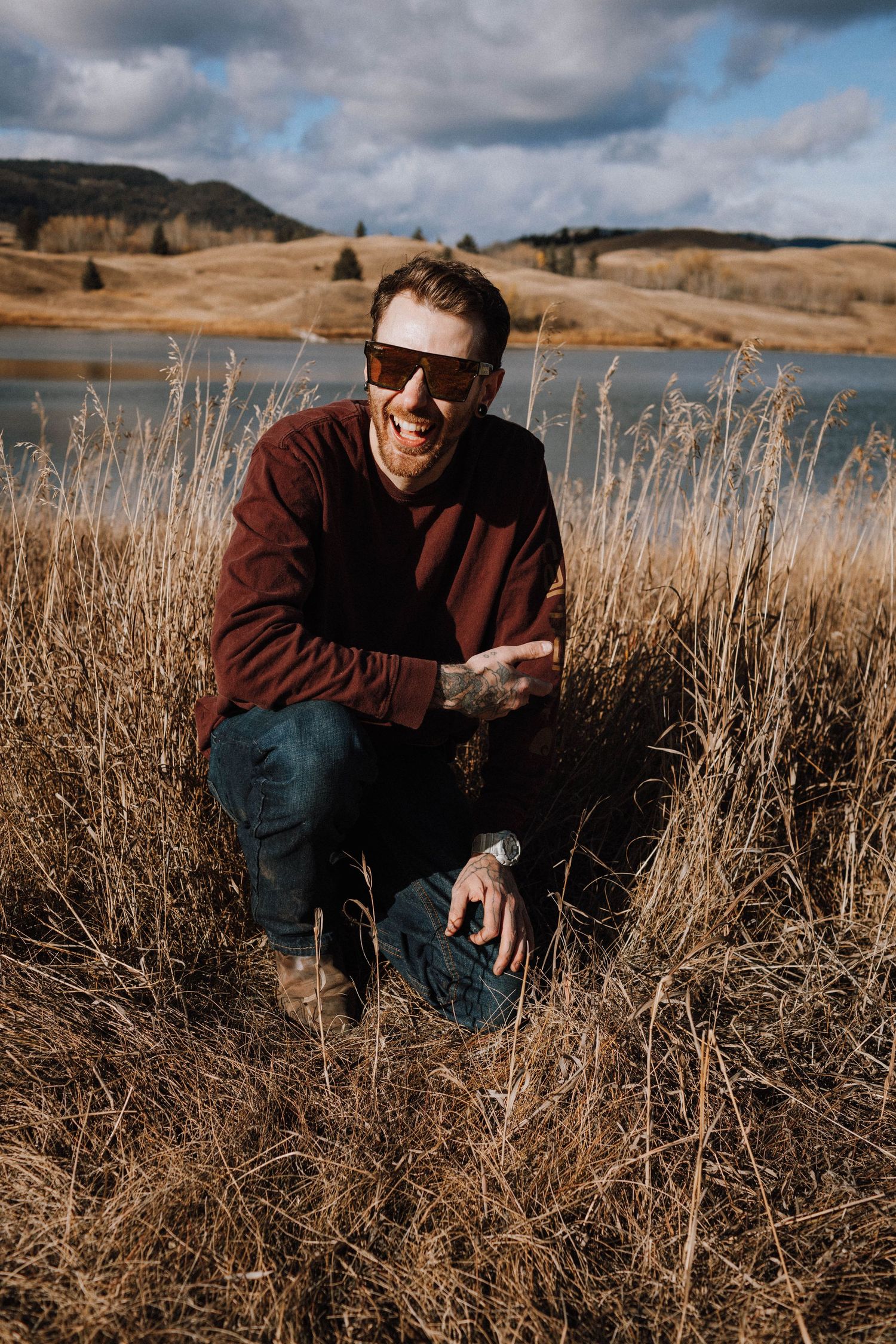 Someone in a brown sweater and sunglasses crouches in tall grass by a lake on a cloudy autumn day.
