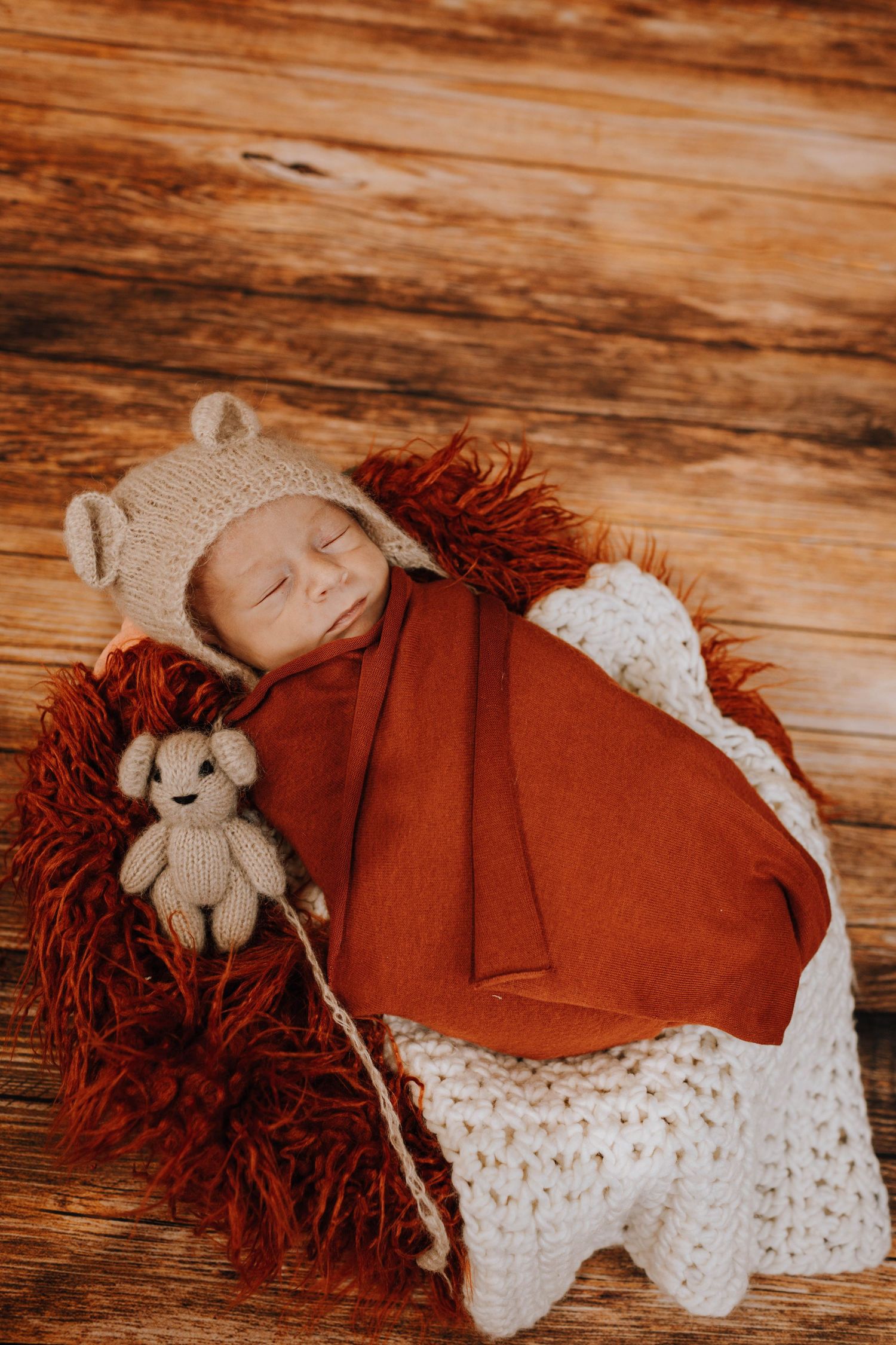 A sleeping baby in a bear hat lies wrapped in a red blanket on red fur with a knitted toy companion on wooden floor.