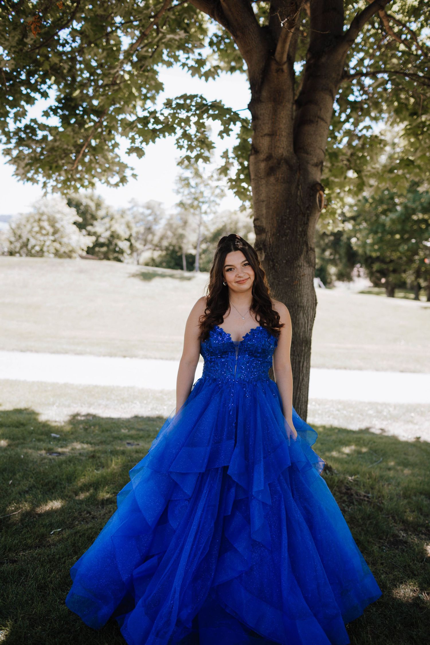 A royal blue tulle ballgown sparkles in the sunlight under a large tree in a park setting.