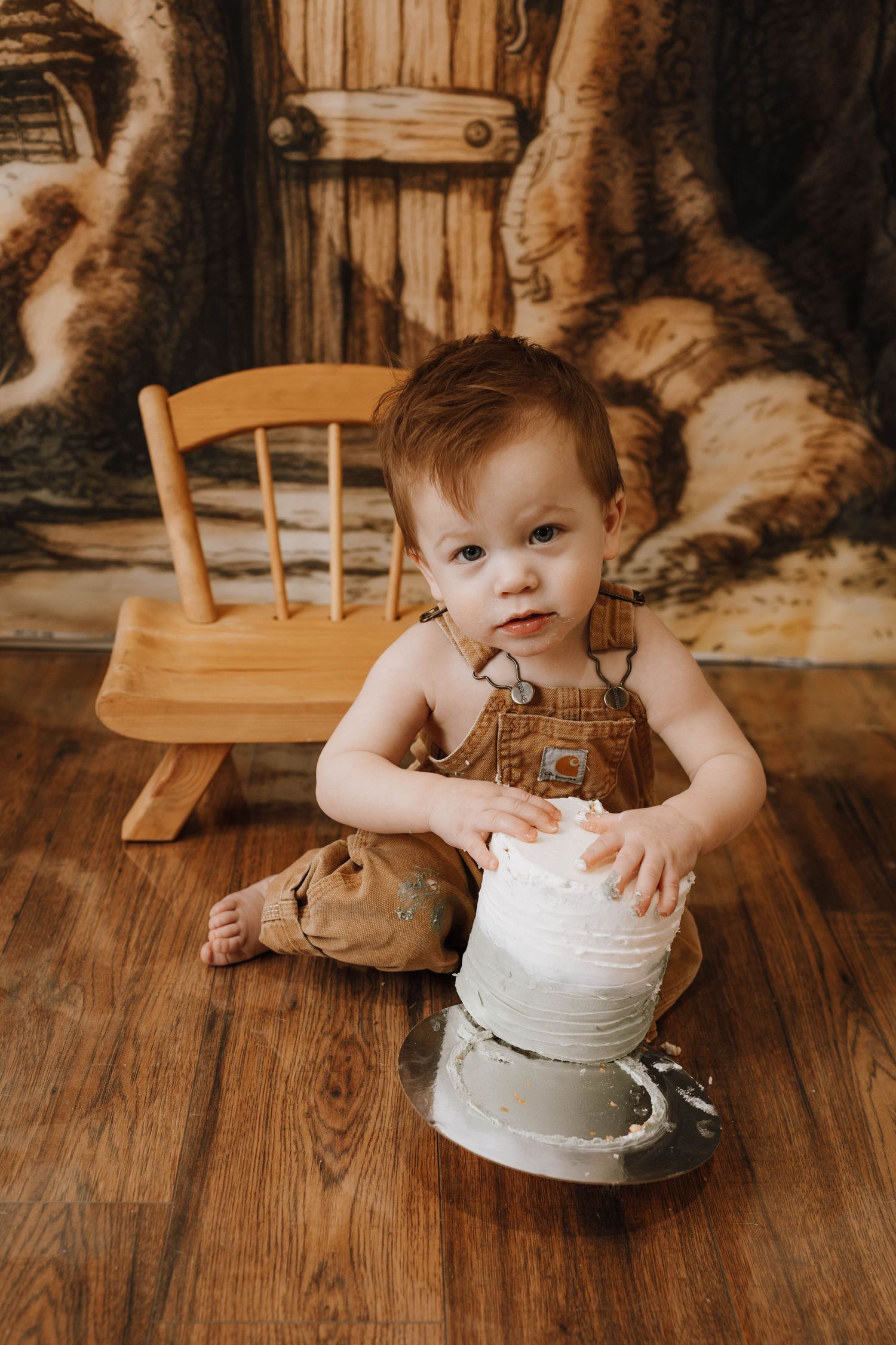 Small child in brown overalls plays with a white pot on wooden floor.