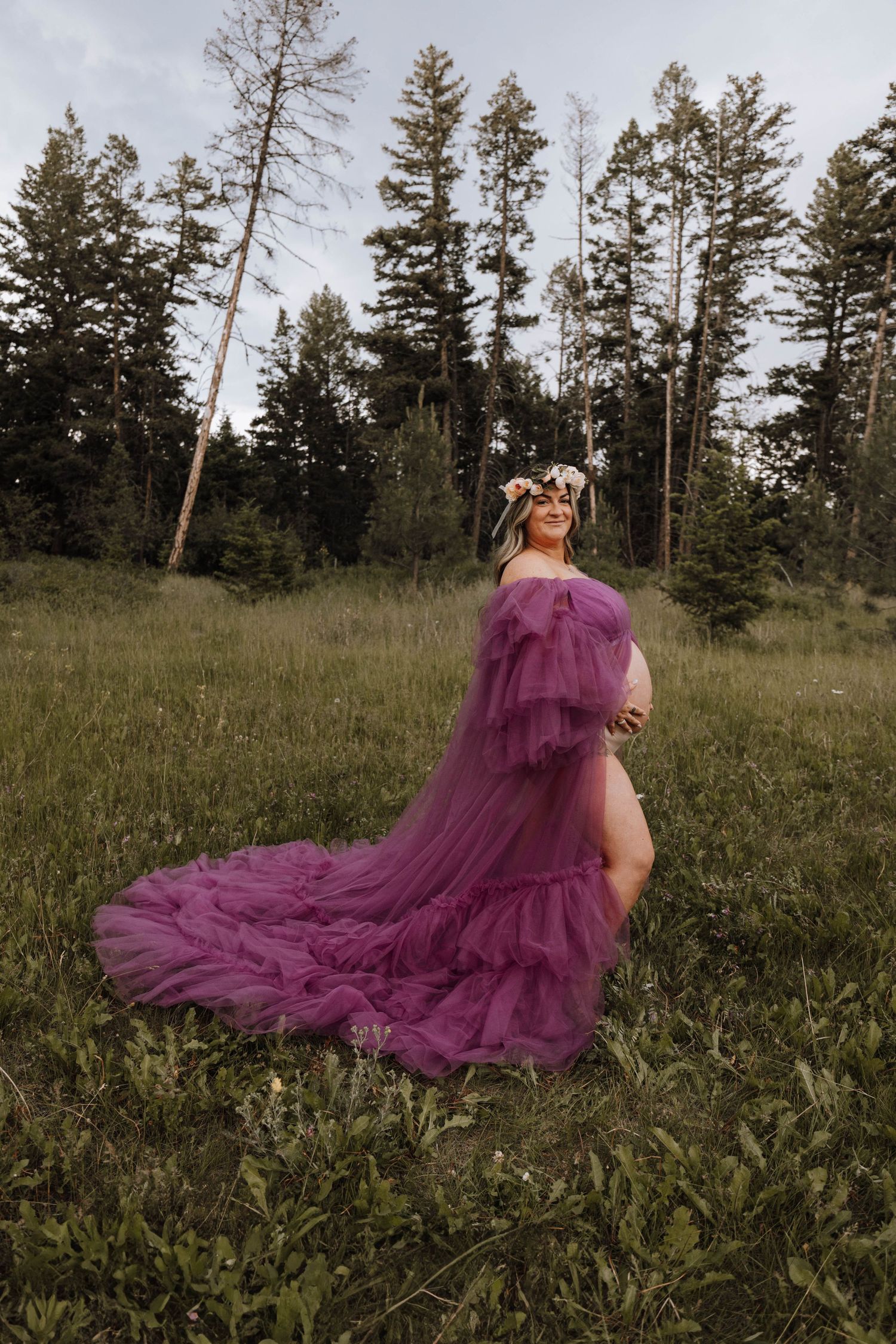 A person in a flowing purple tulle gown poses dramatically in a grassy meadow surrounded by tall evergreen trees.