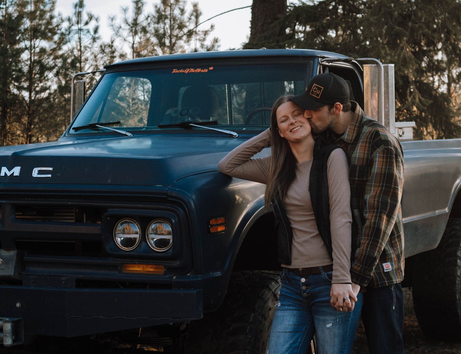 A couple poses against a vintage GMC pickup truck in a wooded setting.