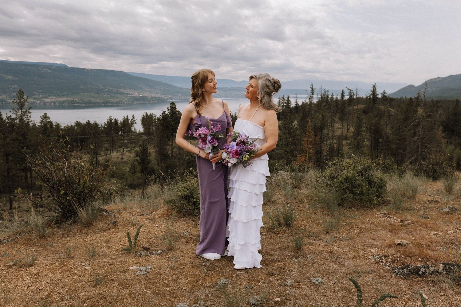 Two people in formal dresses stand together on a hilltop overlooking a scenic lake and mountains on a cloudy day.