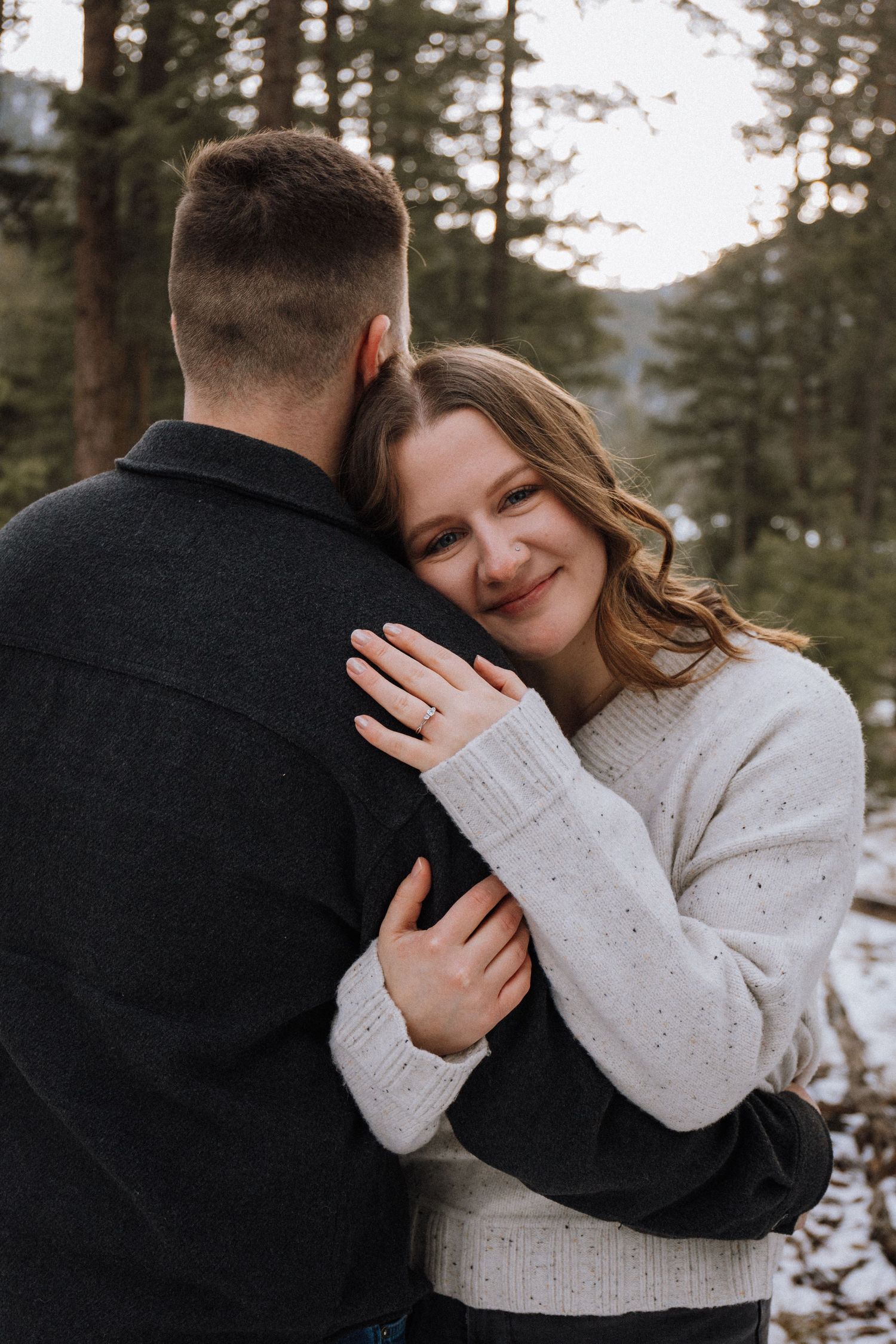 A smiling woman in a white sweater embraces a man in a dark jacket outdoors in a snowy forest setting.