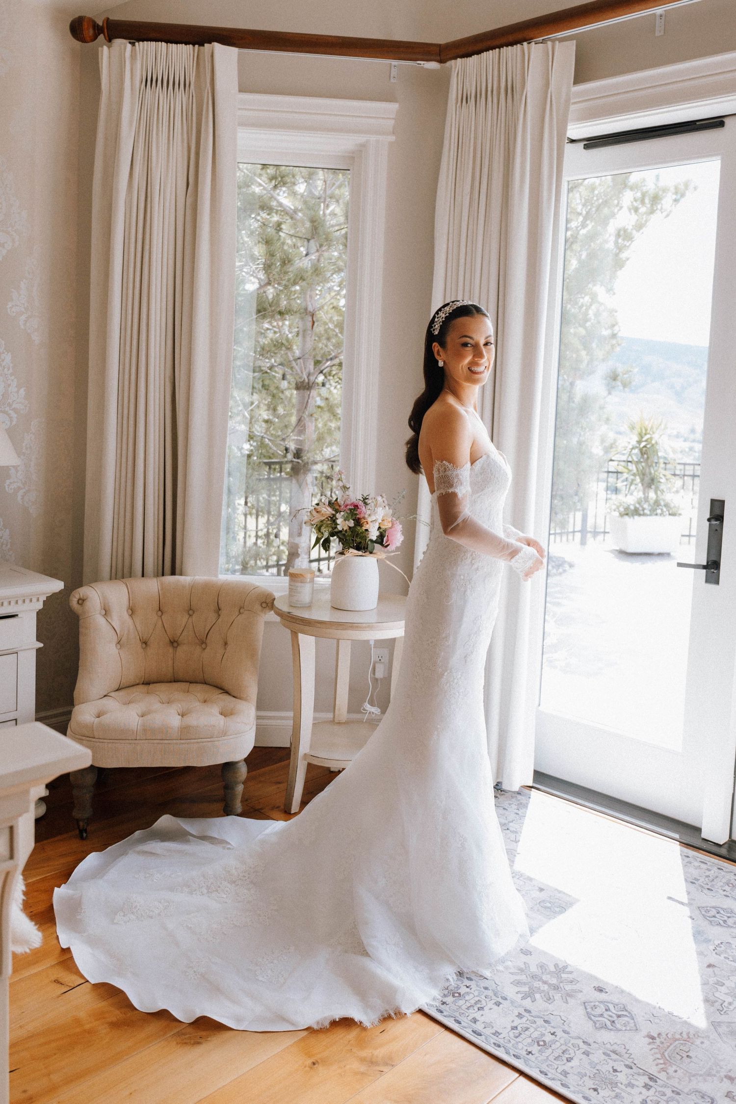 A bride in an elegant white lace wedding dress with a long train stands by sunlit windows in an elegant room with white curtains.