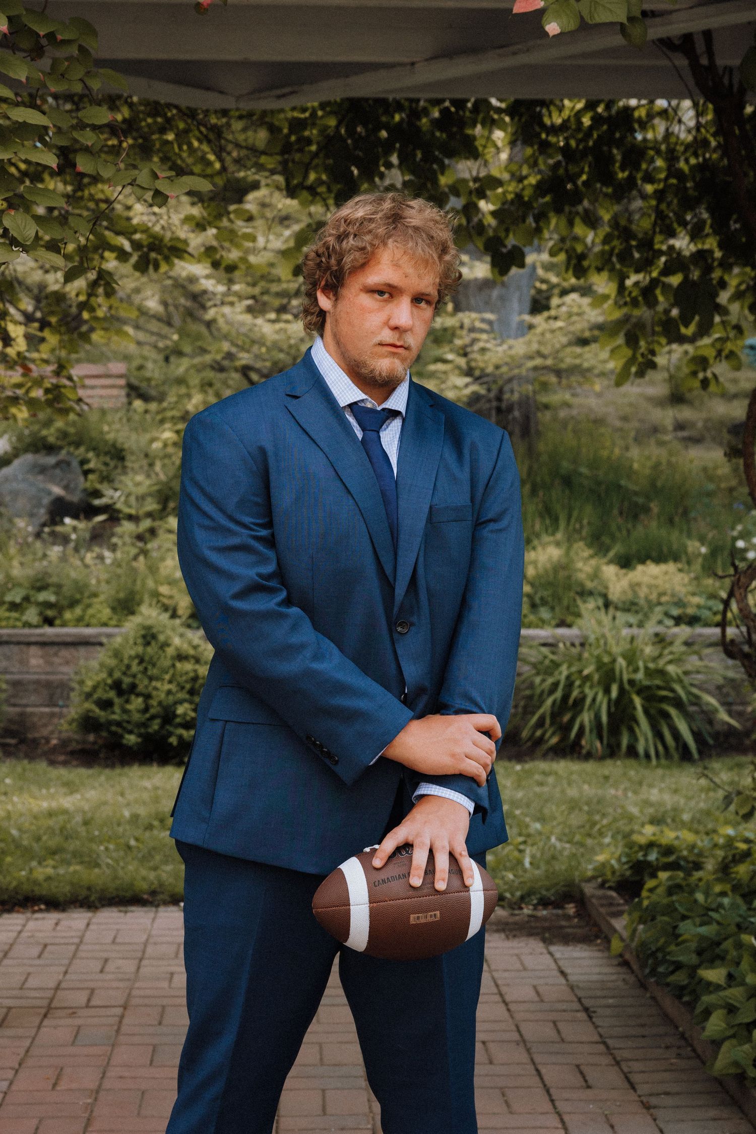 Young man in blue suit holding football poses in lush garden setting for formal portrait photo.