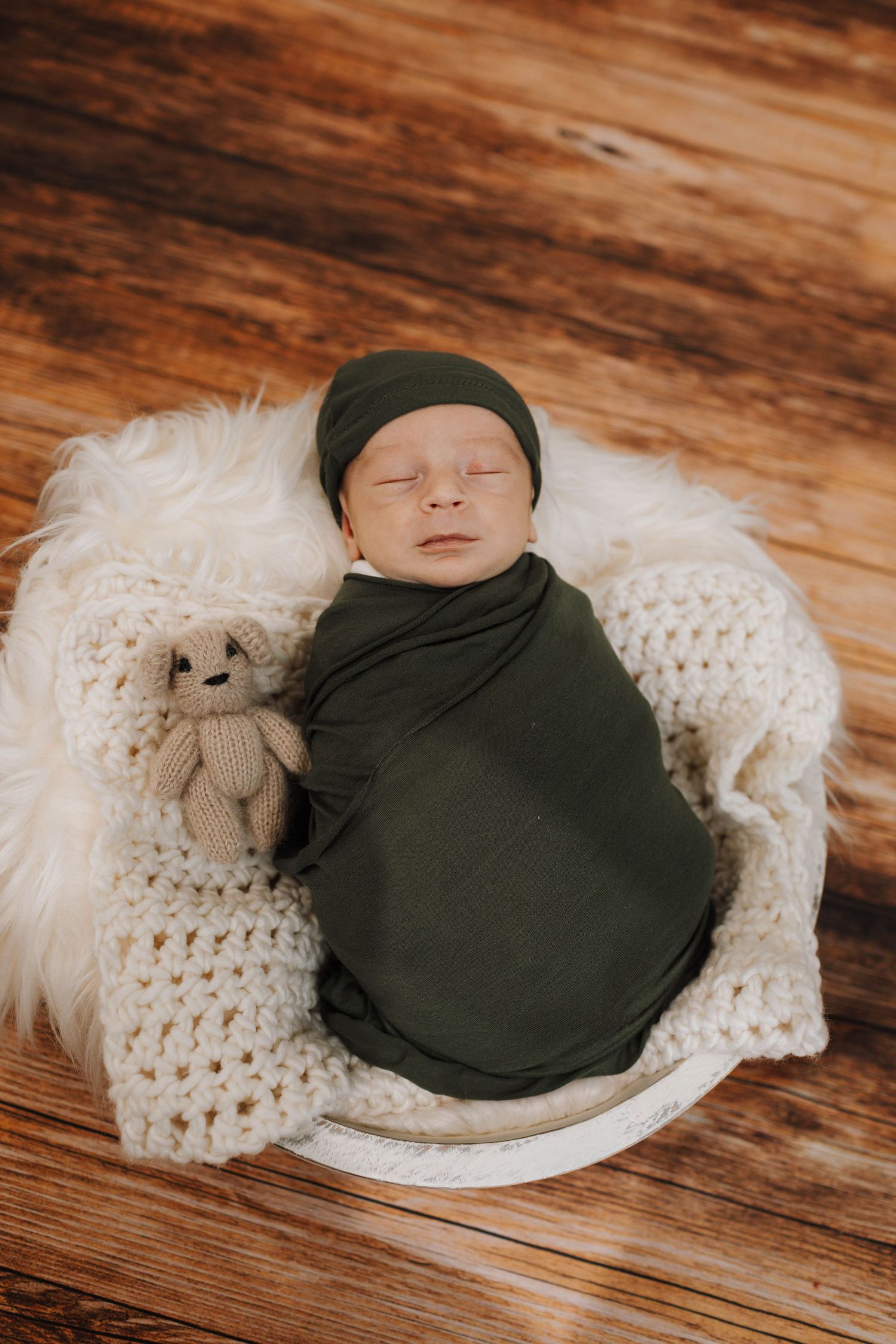 Newborn baby wrapped in dark green swaddle rests on white knit blanket with small plush toy on wooden floor.