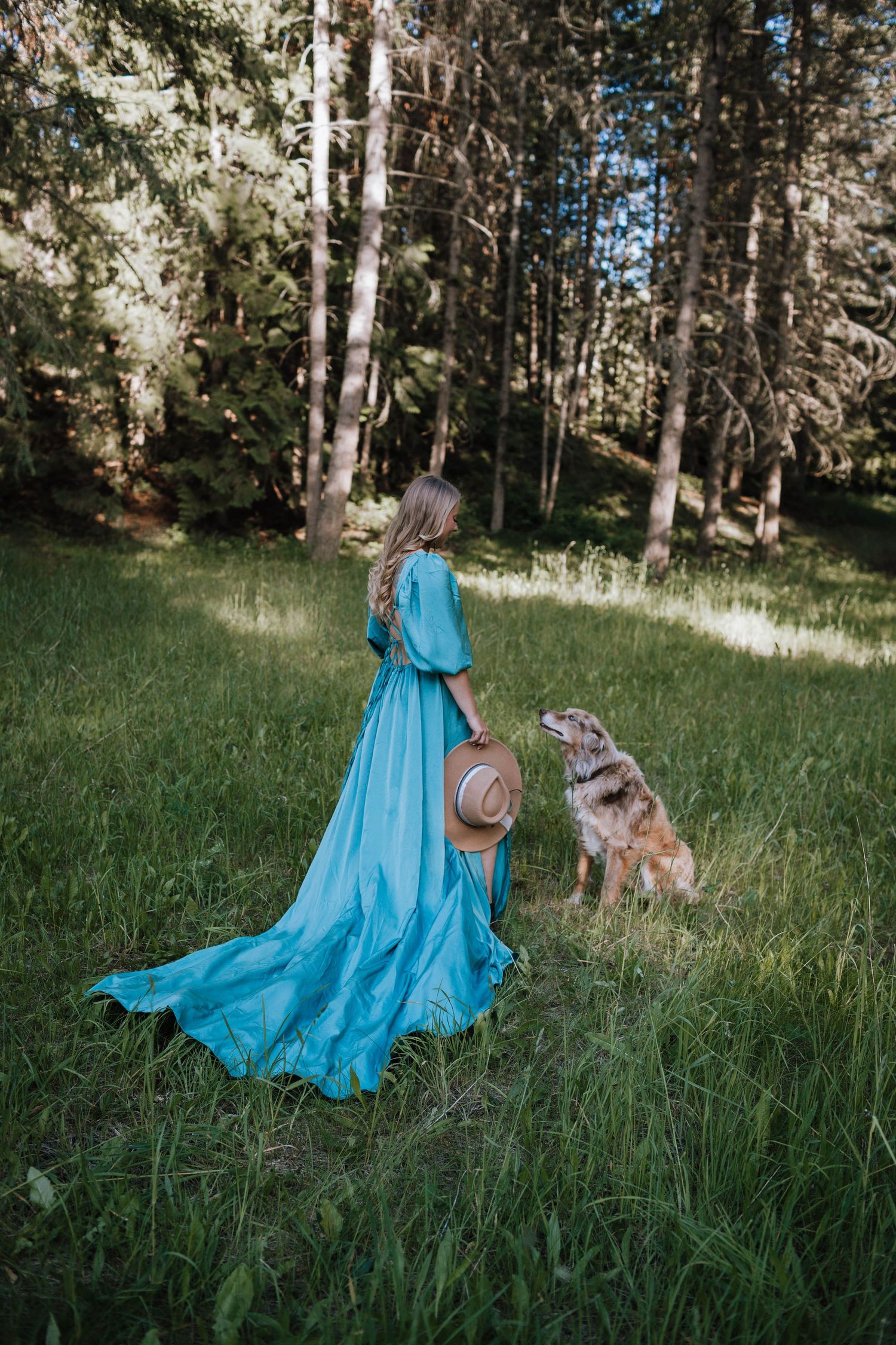Flowing turquoise dress trails behind as person walks through forest with dog companion.