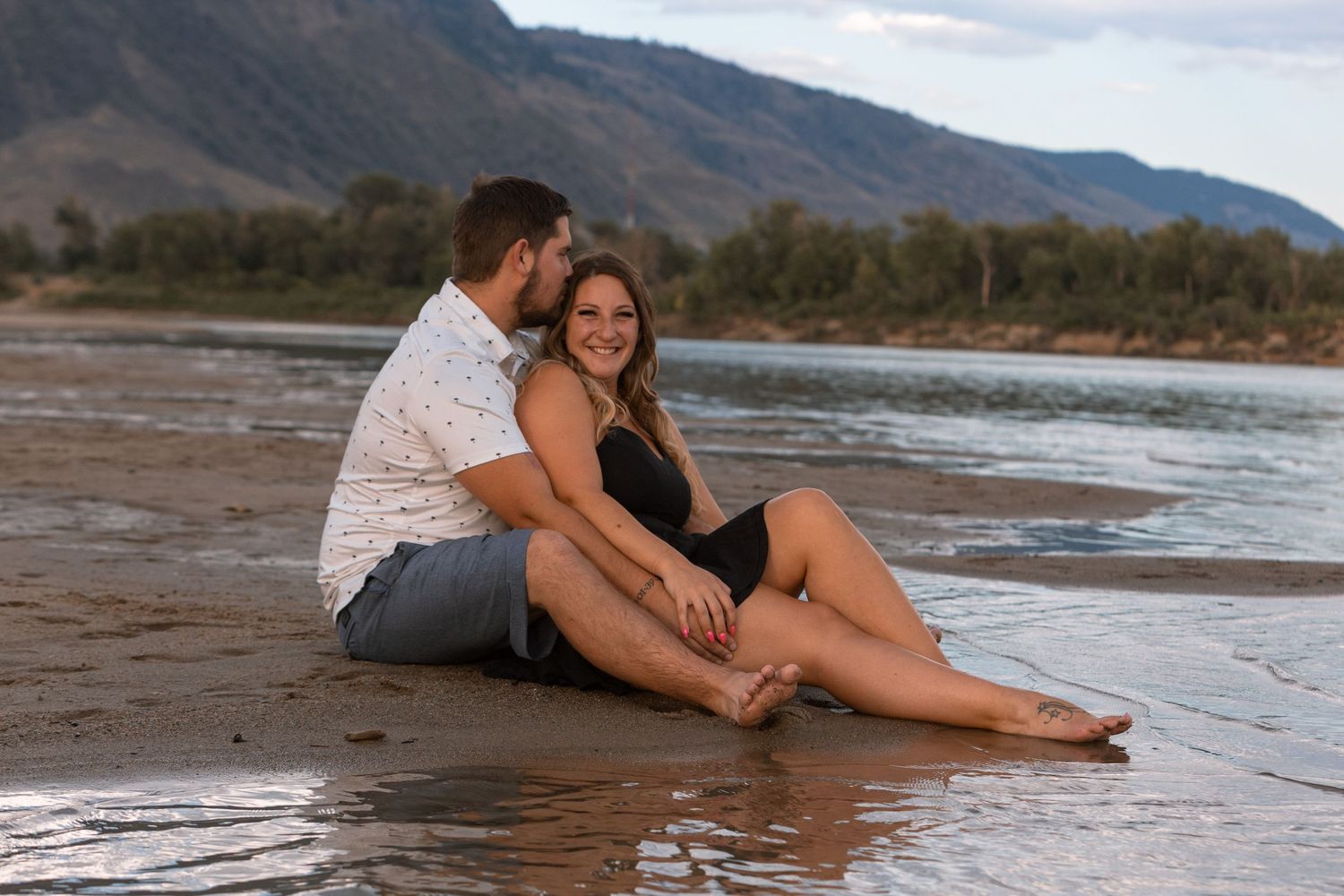 A romantic couple sitting together on the shoreline with mountains in the background.