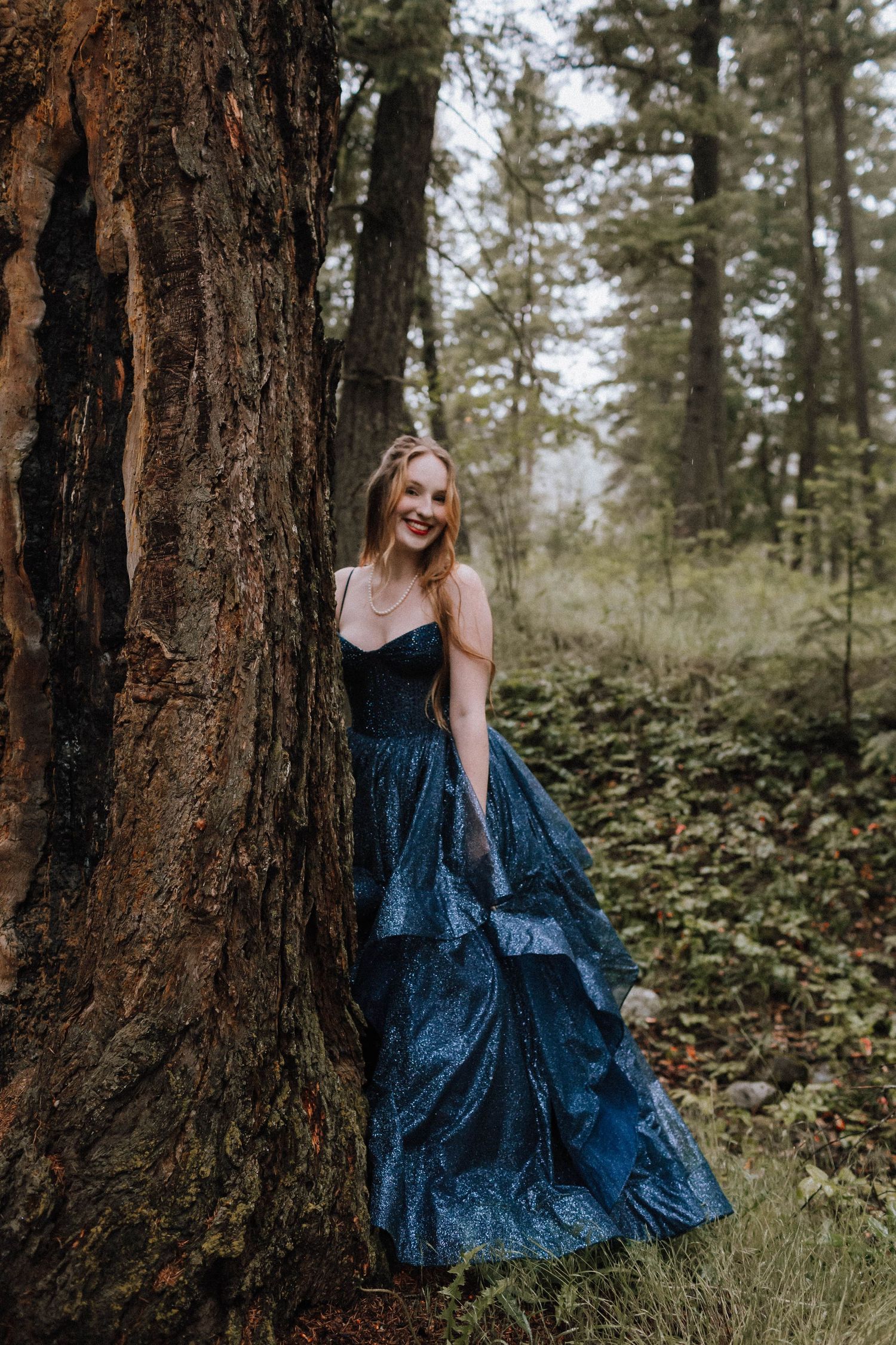 A sparkly navy blue ballgown contrasts beautifully against a large tree trunk in the forest.