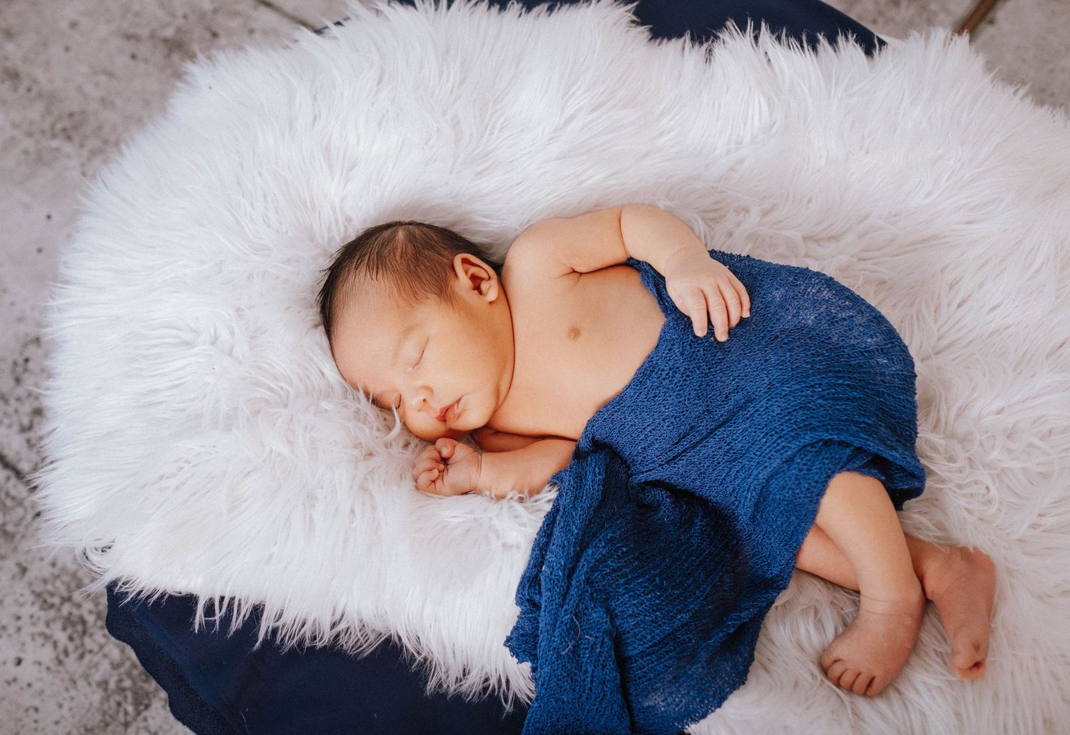 Sleeping newborn wrapped in blue blanket on white furry rug.