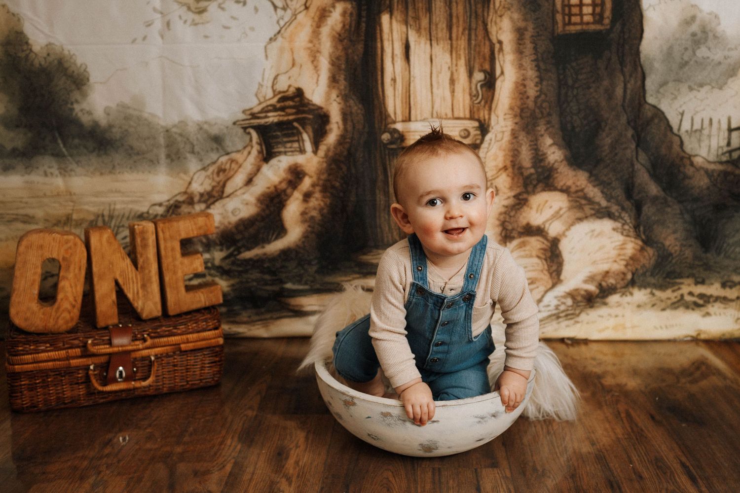Happy baby in blue overalls sitting in white bowl against fairy tale forest backdrop.