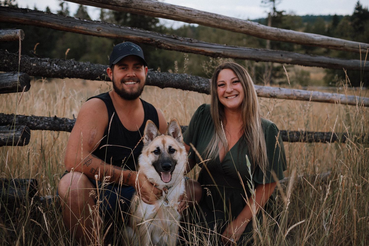 A couple poses with their German Shepherd dog near a rustic wooden fence.