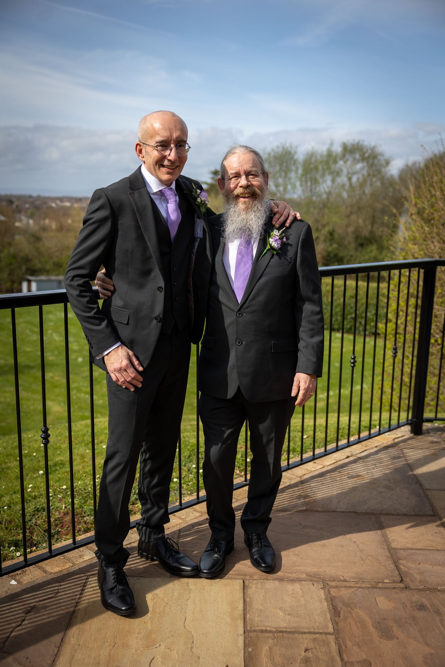 Two wedding guests in black suits with purple accessories standing on an outdoor terrace.