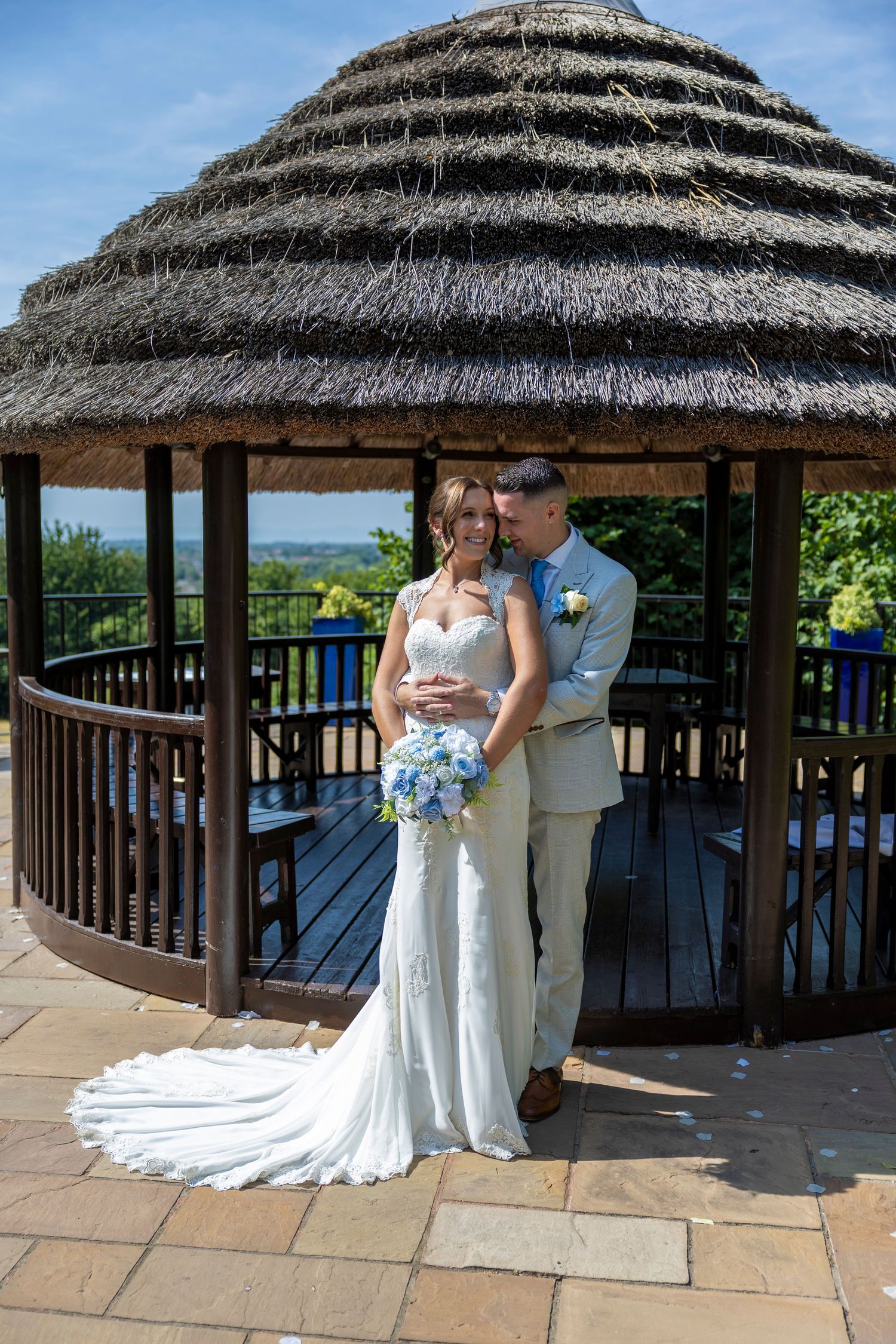 Bride and groom embracing under a thatched gazebo, bride holding blue bouquet, on a sunny wedding day.