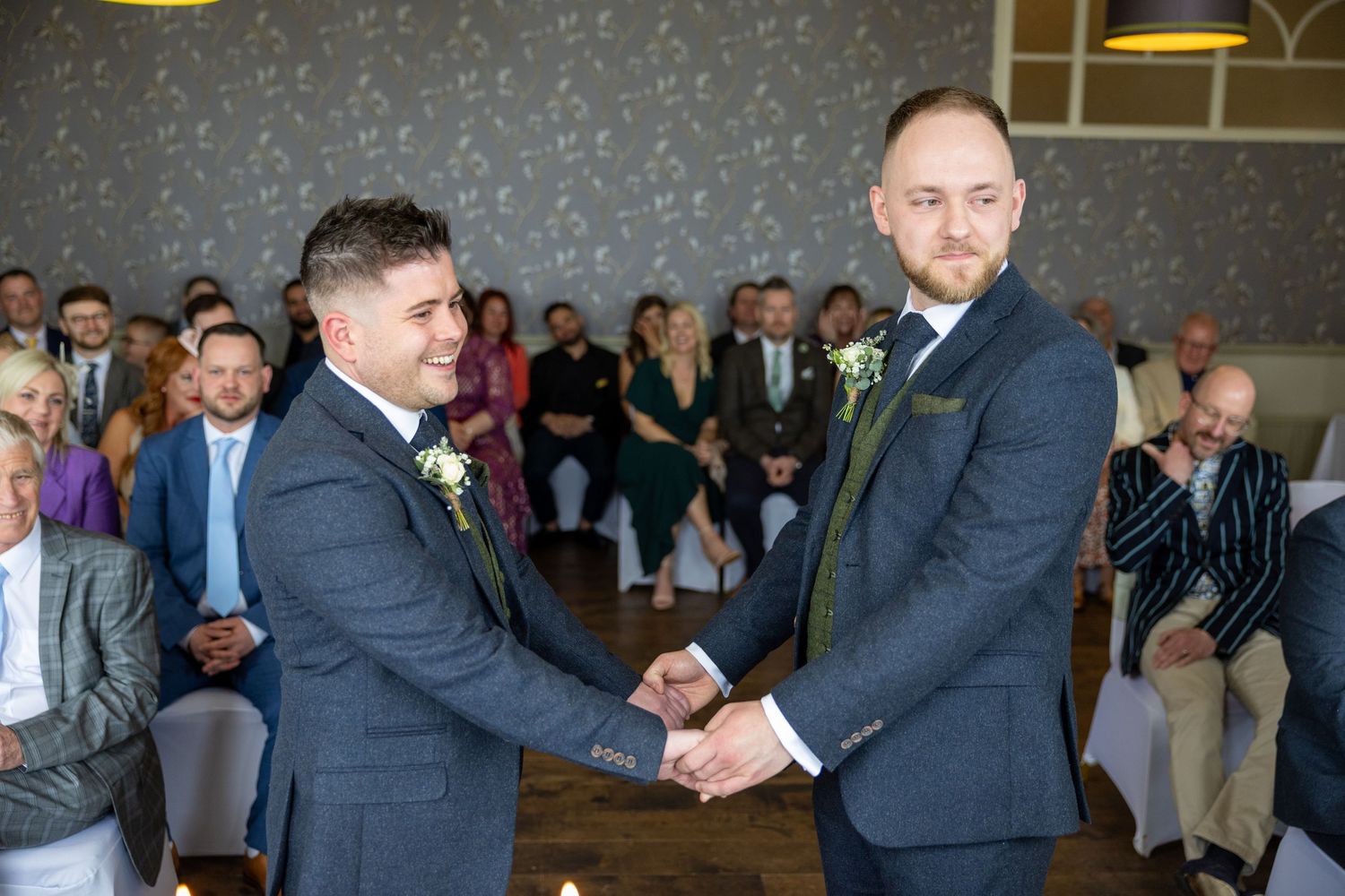 Two men in suits shake hands during a wedding ceremony with guests watching in the background.