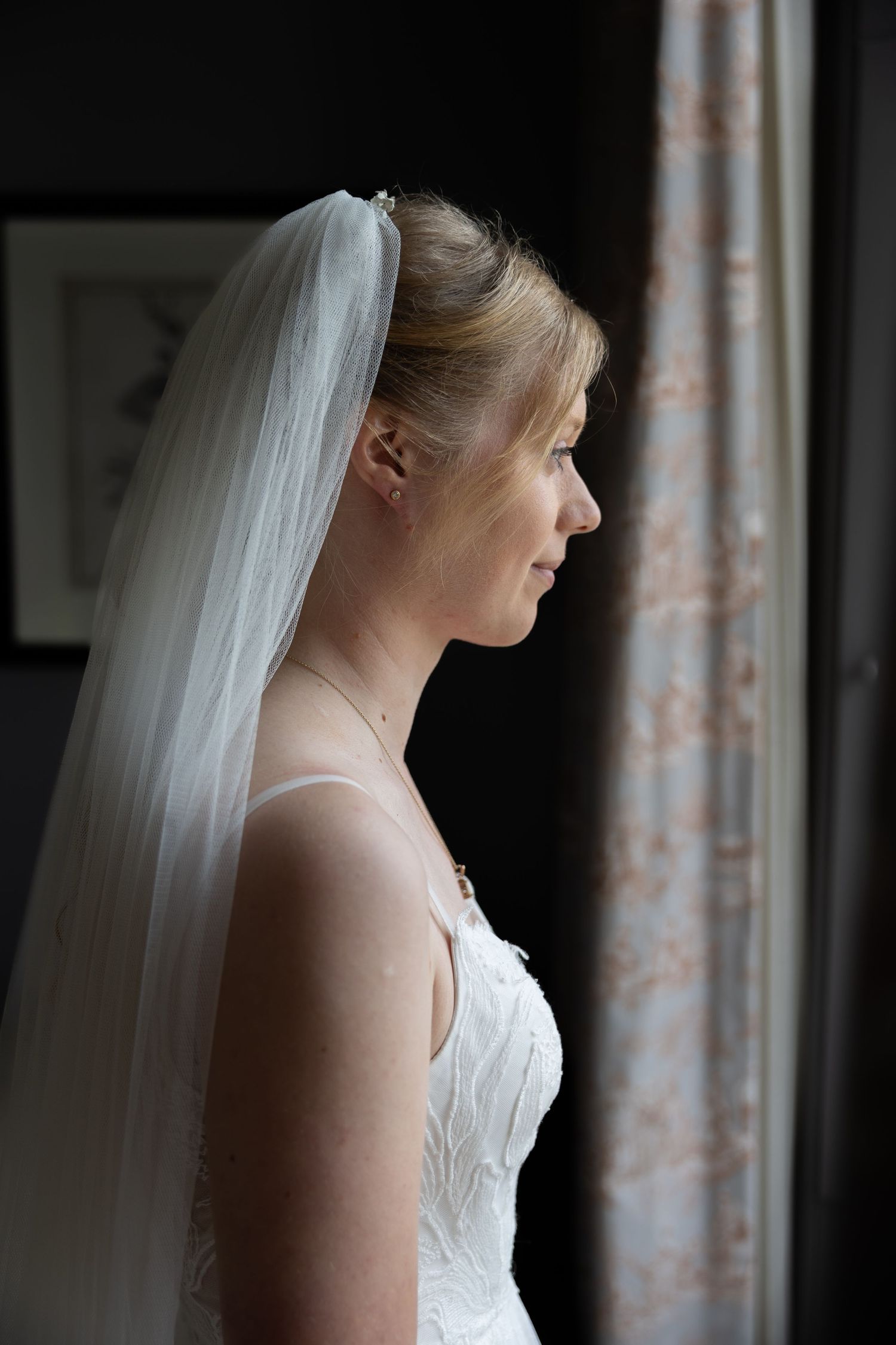 Side profile of a bride wearing a white wedding dress and long veil standing by a window in natural light.