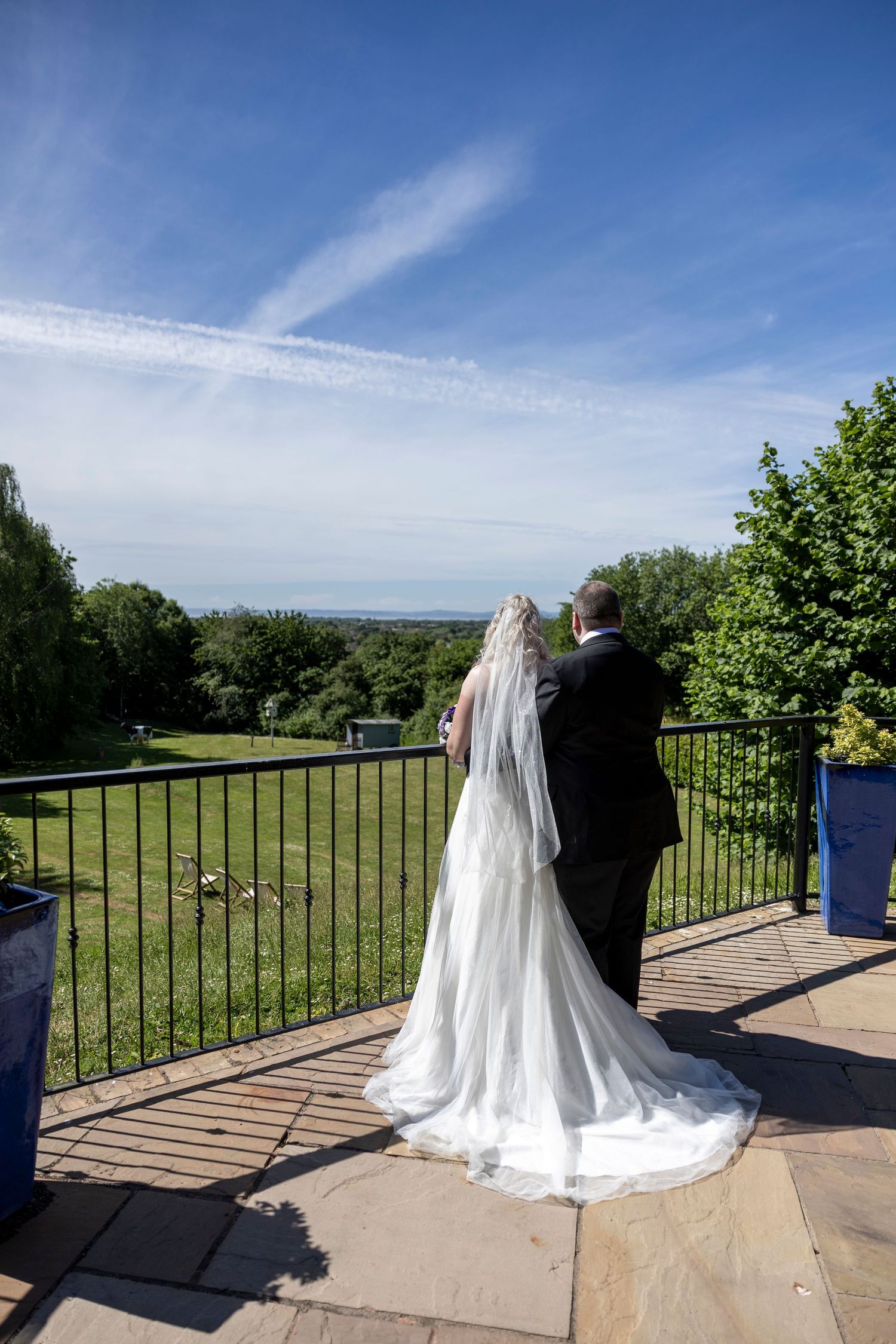 Bride and groom stand on a terrace overlooking lush green gardens under a bright blue sky on their wedding day.