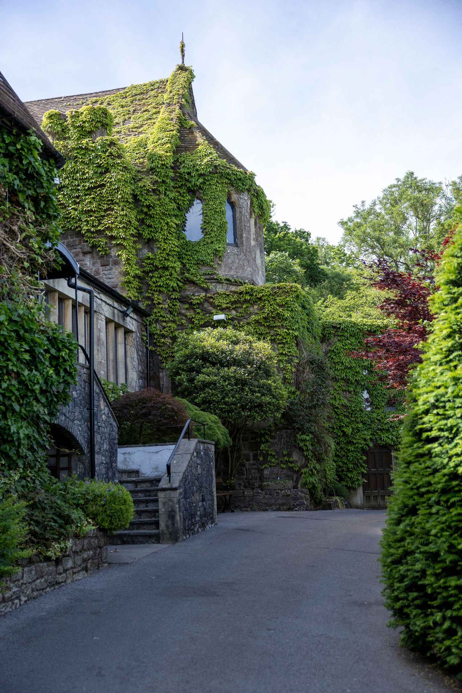 Ivy-covered stone building with lush green gardens, stone steps, and a winding pathway in a serene countryside setting.