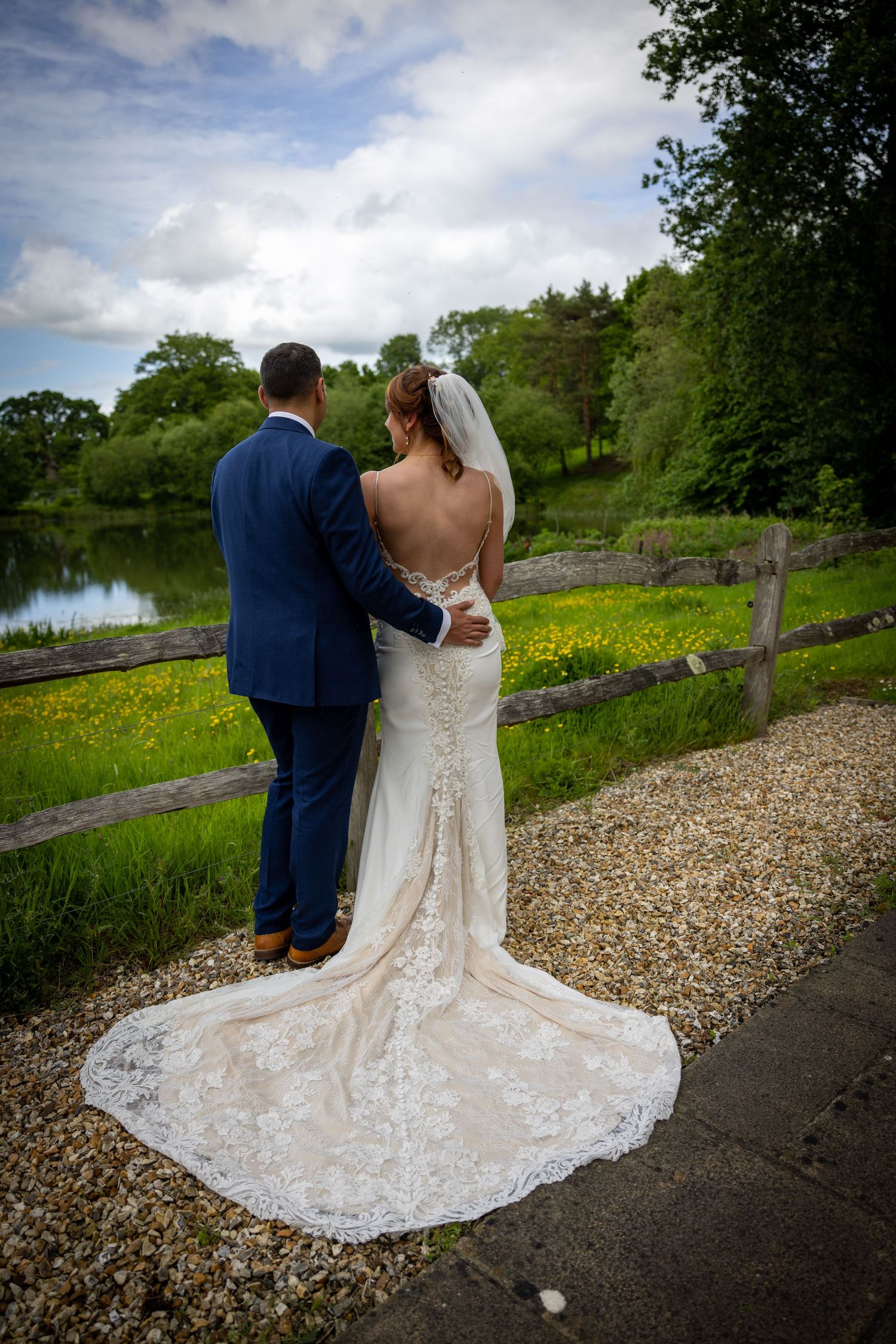 A bride and groom stand together by a scenic lake with lush greenery and wildflowers.