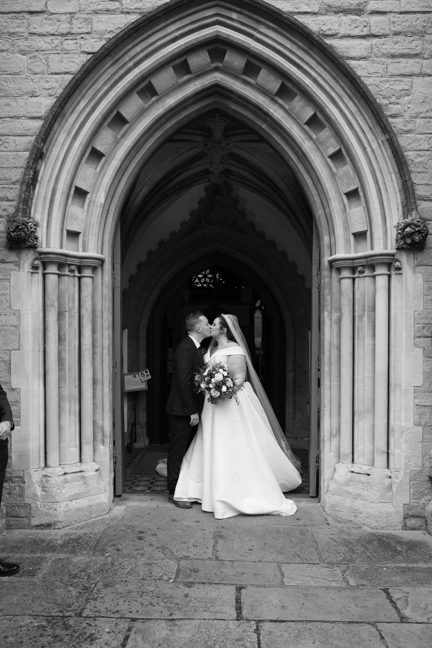 A black and white photo of newlyweds framed by a gothic stone church archway.