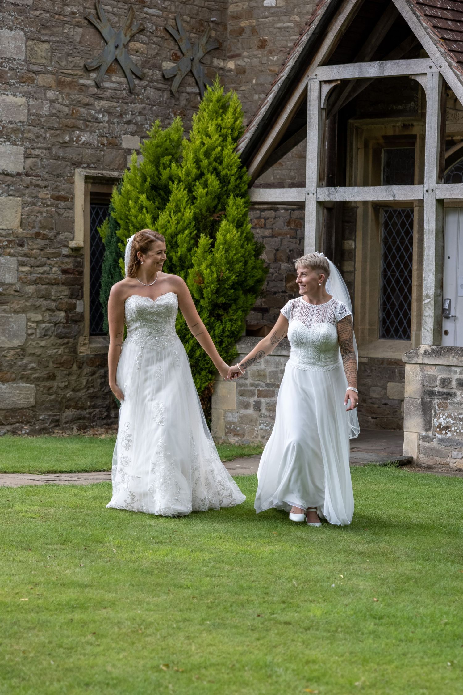 Two people in elegant white dresses holding hands while walking on a manicured lawn outside a stone church.