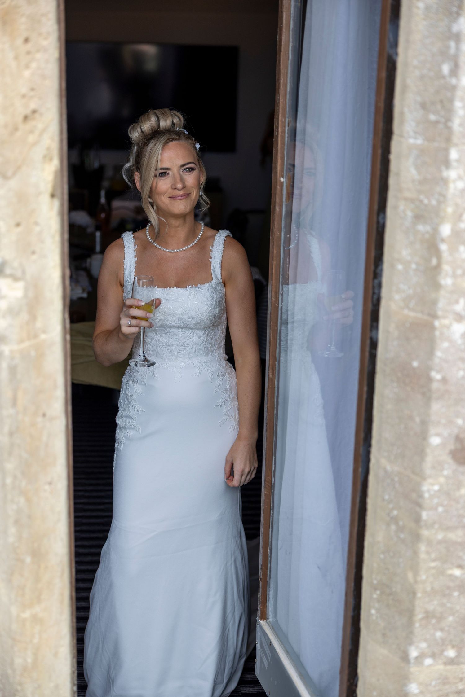 Bride in white lace gown holding champagne glass standing in stone doorway with curtains.