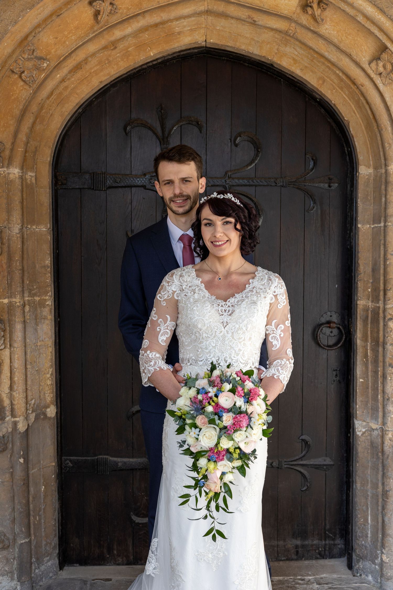 Wedding portrait in front of an ornate wooden church door with stone archway.
