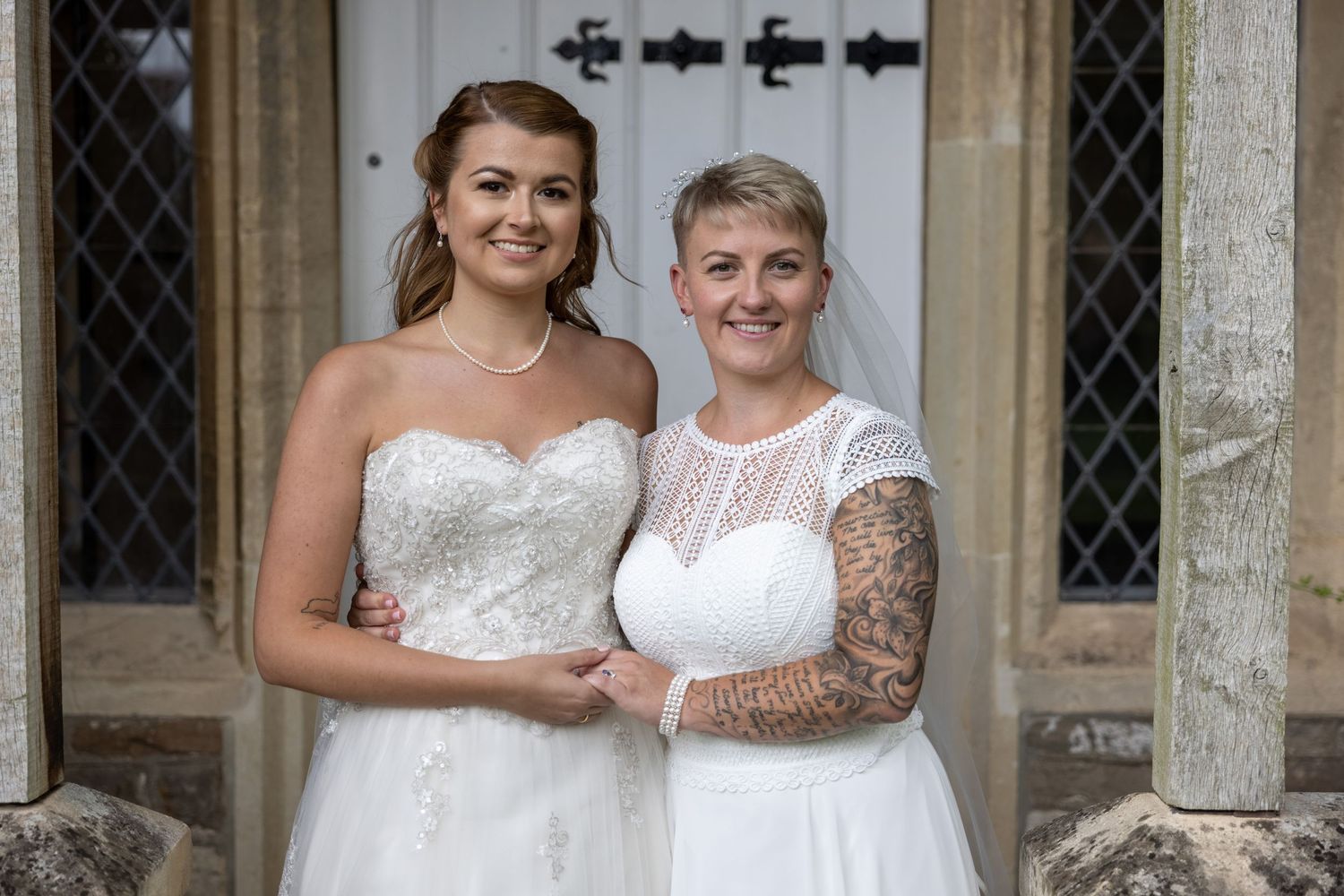 Two people in white wedding dresses pose together in front of a church entrance with stone columns and windows.