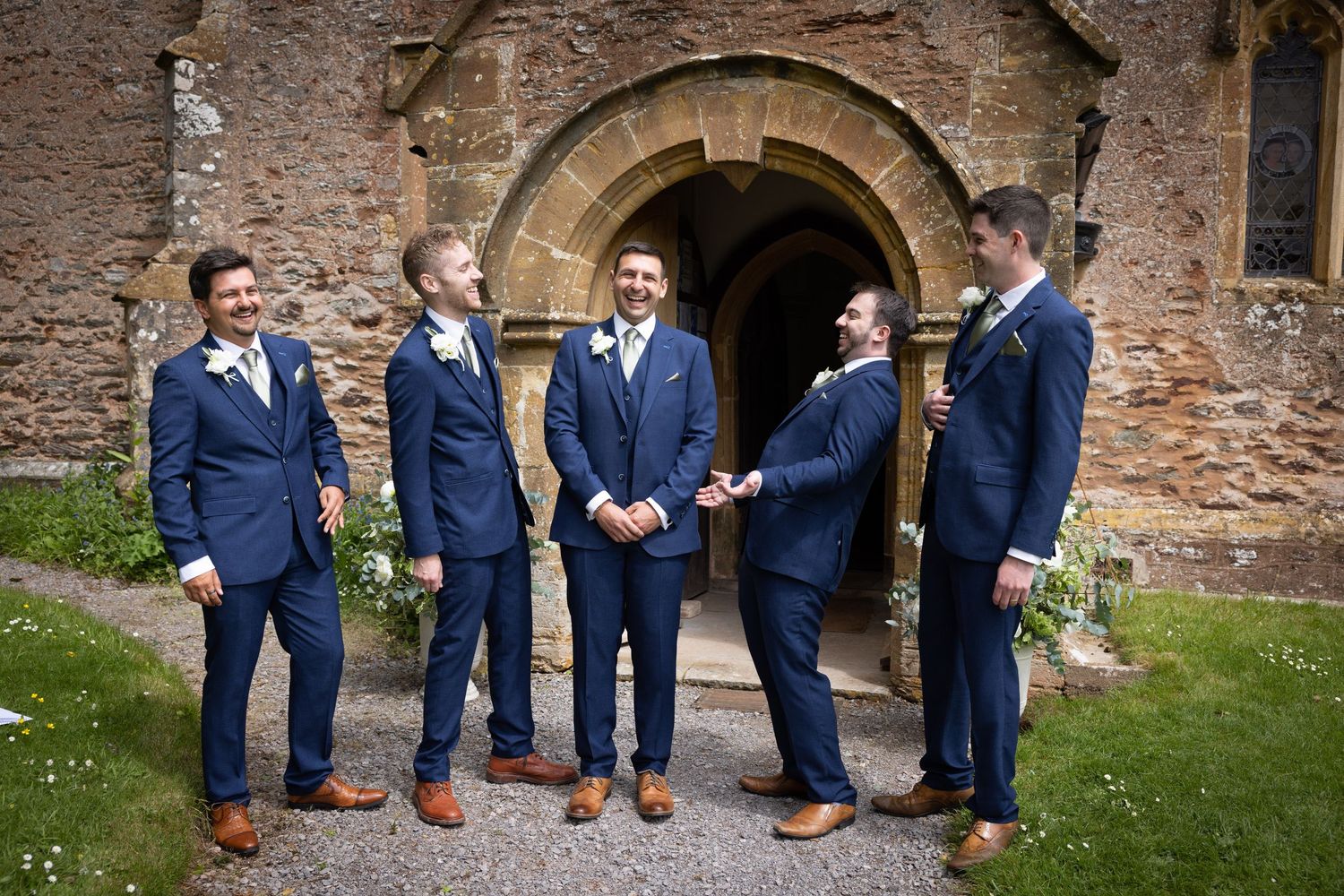 Five groomsmen in matching navy suits and brown shoes stand together laughing in front of a stone archway.
