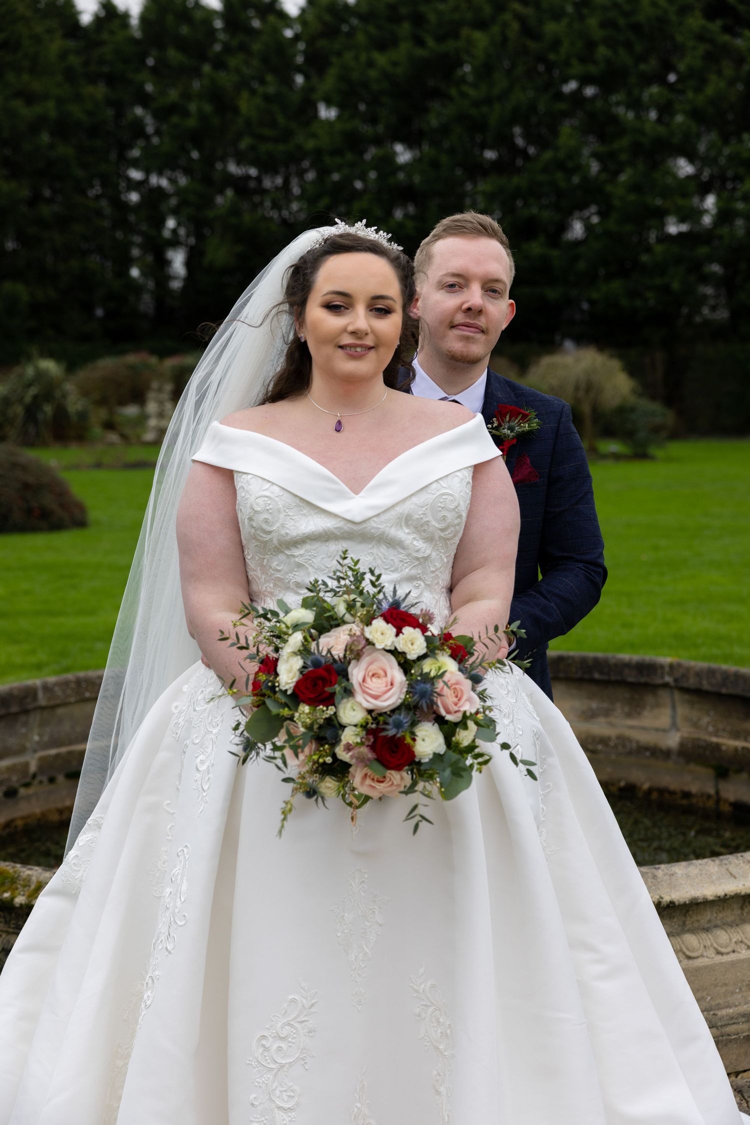 A wedding couple poses in a garden setting with a beautiful floral bouquet.