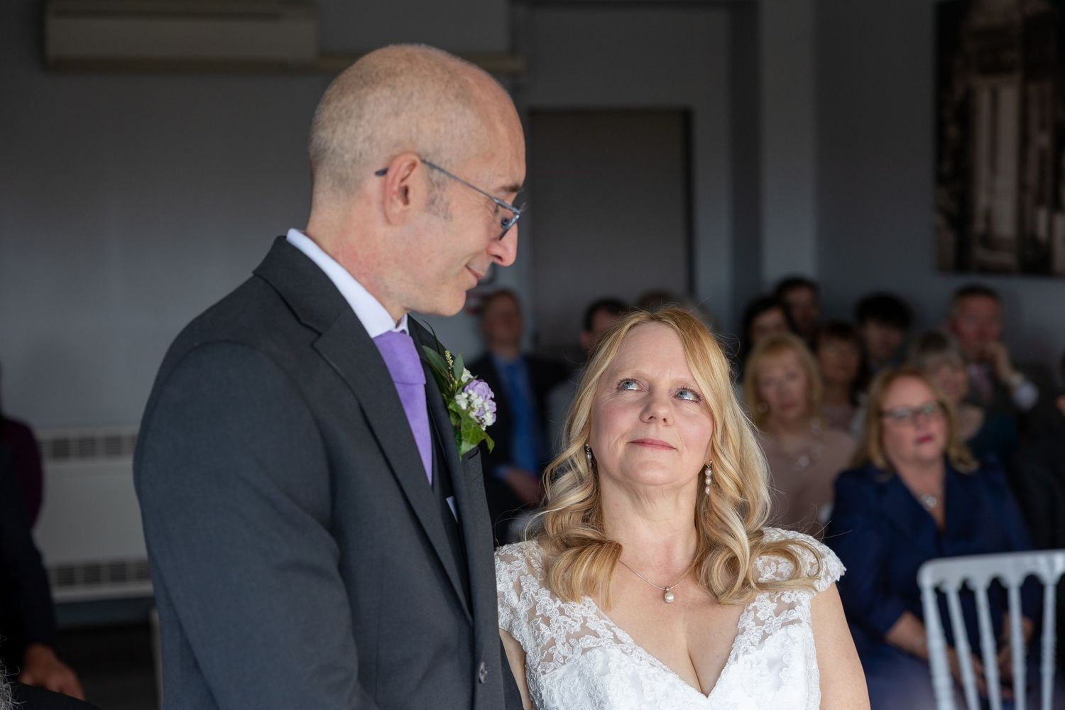 An intimate wedding ceremony moment captured indoors with guests visible in background.