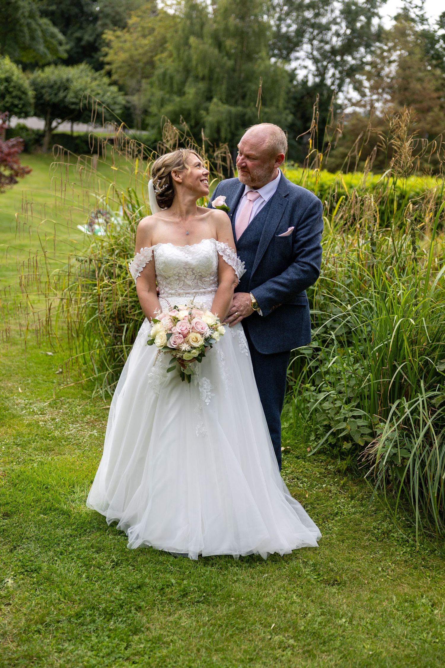 A romantic wedding photo of a couple embracing in a garden with tall ornamental grasses and flowers.