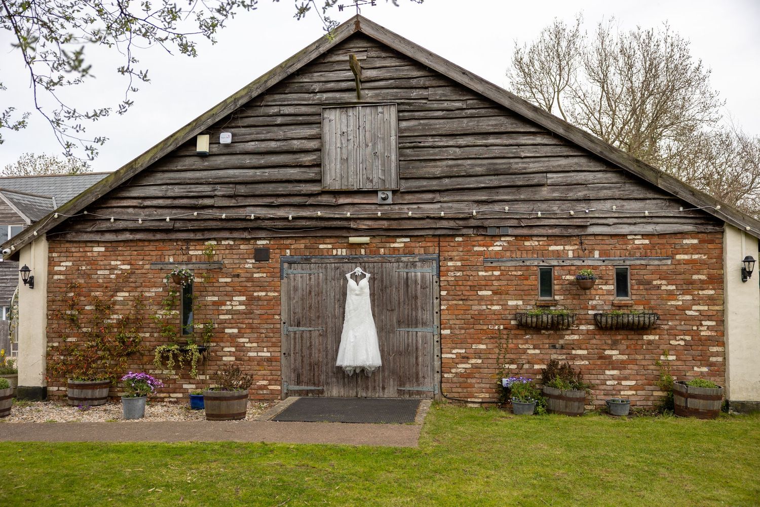 A white wedding dress hangs on a rustic barn door of a brick and wood building with a lawn in front.