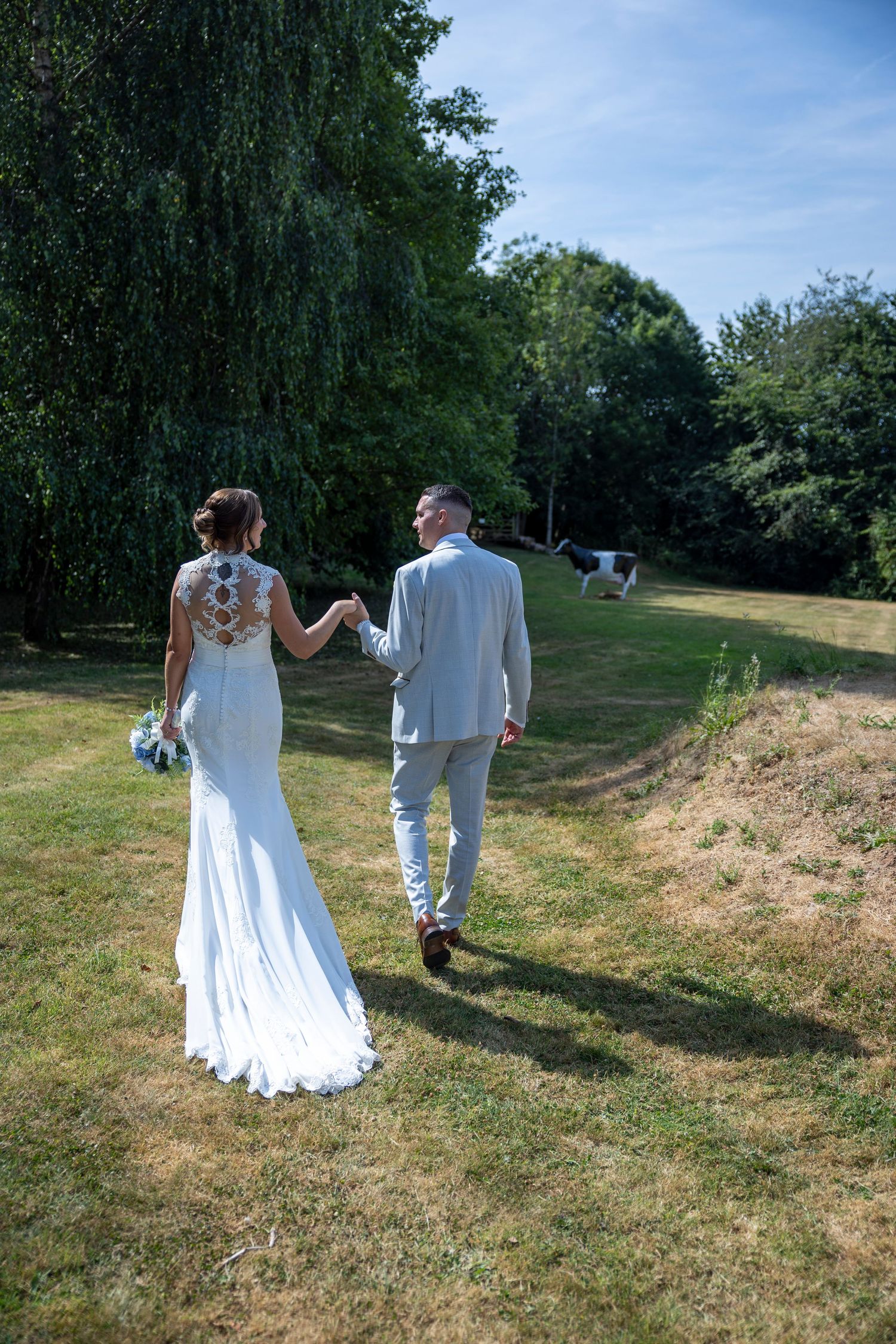 Bride and groom walking hand in hand through a sunny green garden, bride in elegant white lace gown, groom in light gray suit.