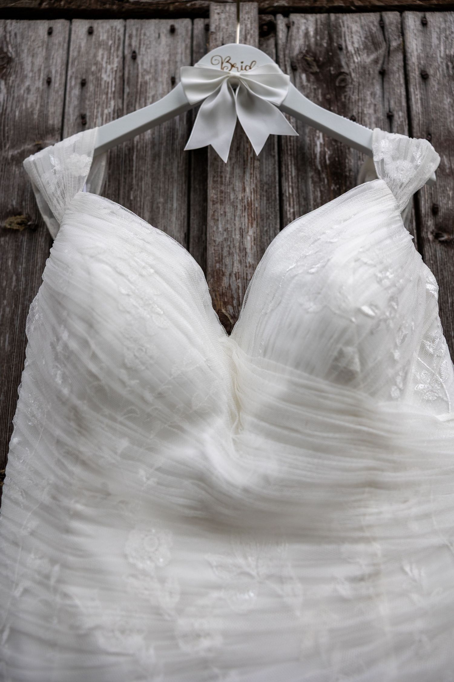 A white wedding dress with sweetheart neckline and gathered tulle fabric hangs on a personalized hanger against rustic wood.