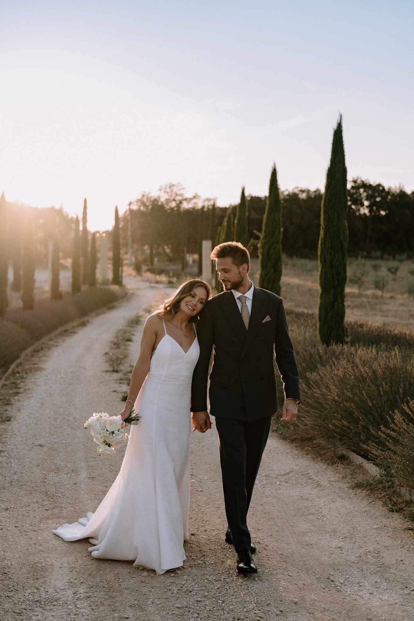 A couple walks hand in hand down a cypress-lined path at sunset in their wedding attire.