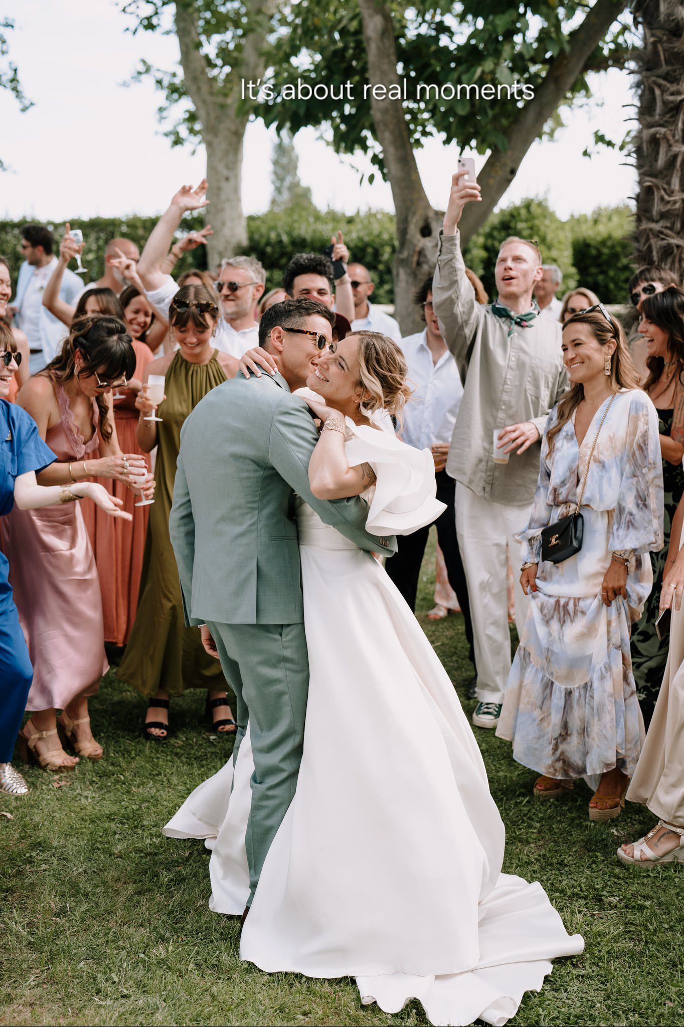 Joyful wedding party celebrates outdoors with dancing and cheering on a grassy lawn under trees.