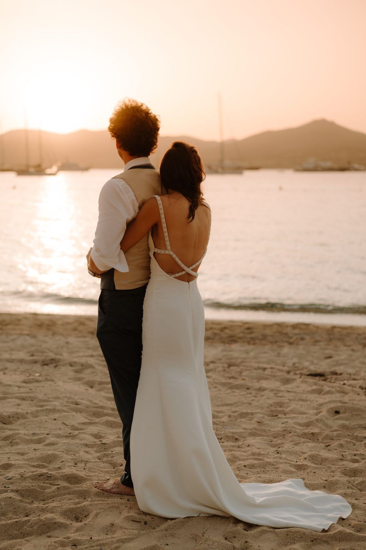 Couple in wedding attire embracing on a beach at sunset.