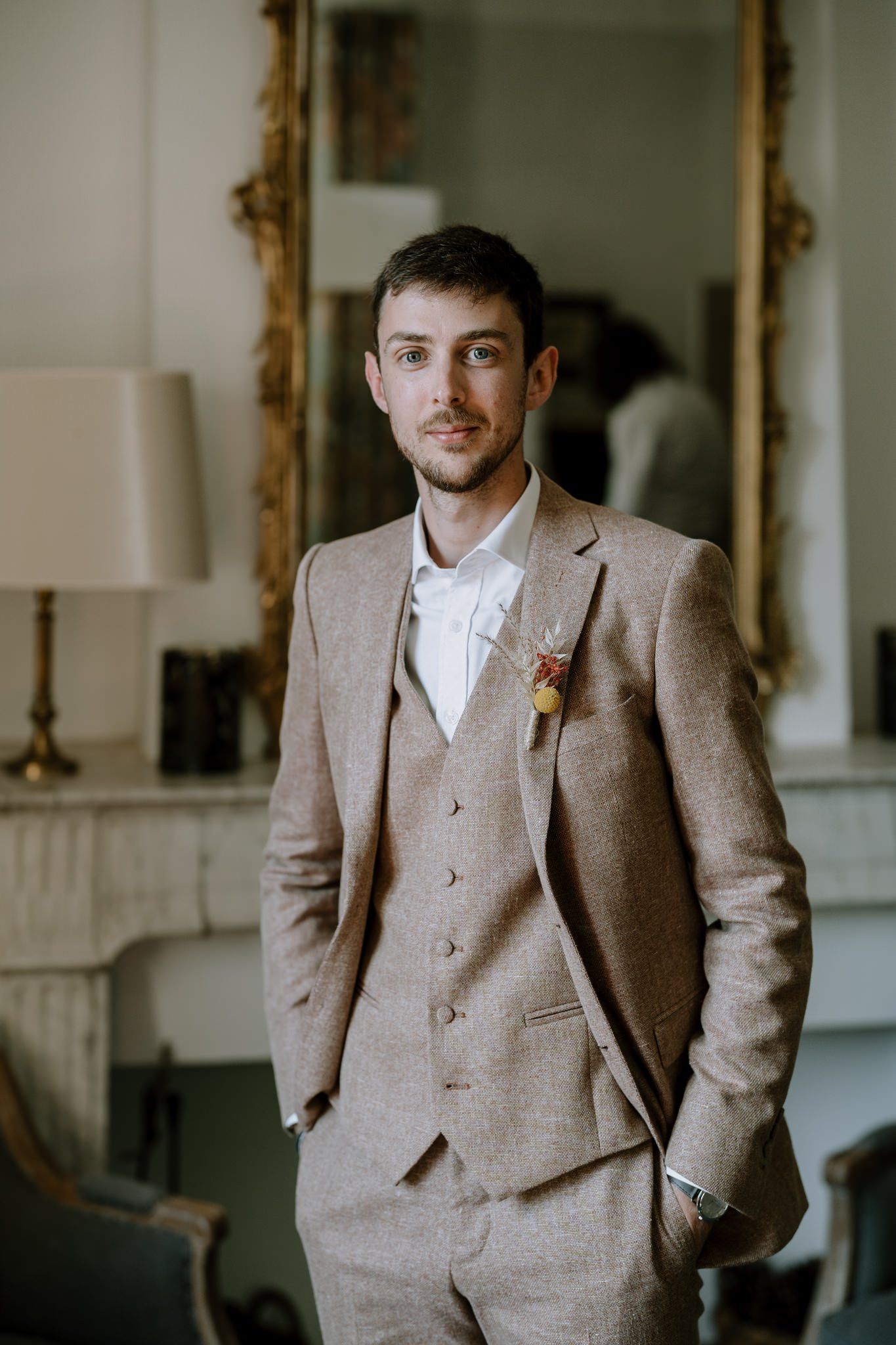 Groom wearing light brown tweed suit standing in elegant interior.