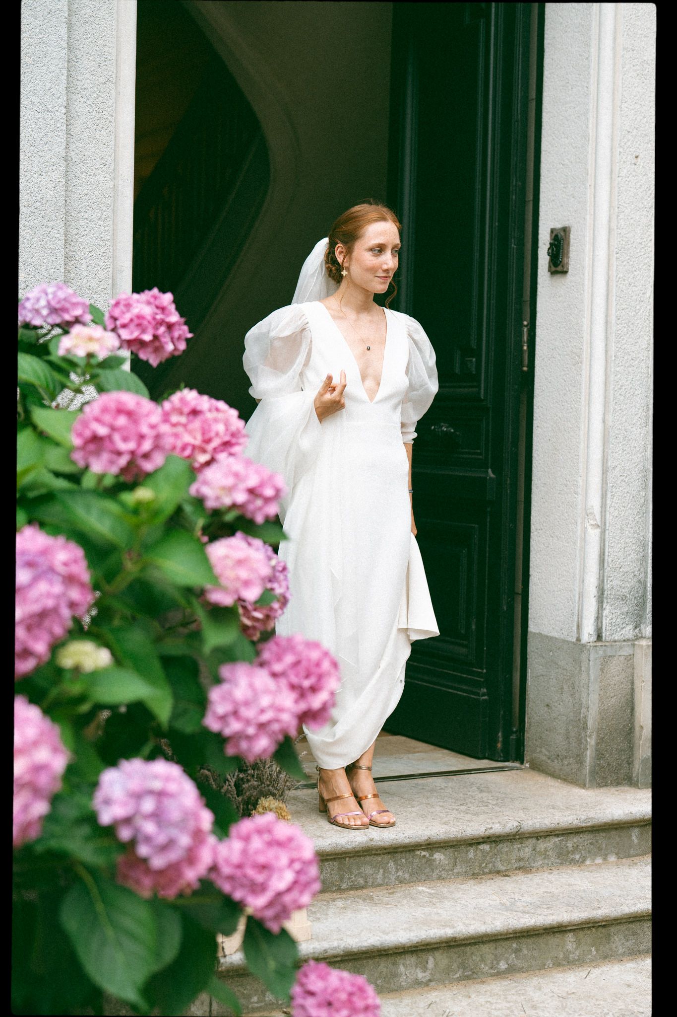 Bride in a flowing white dress poses on stone steps near blooming pink hydrangeas.