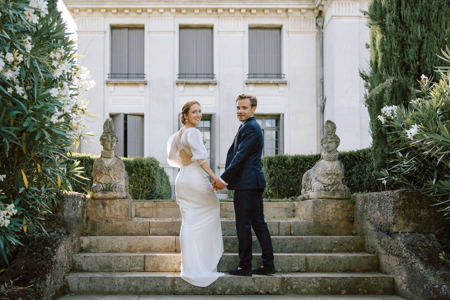 Elegant wedding couple poses on stone steps of a classic mansion with white columns and greenery.