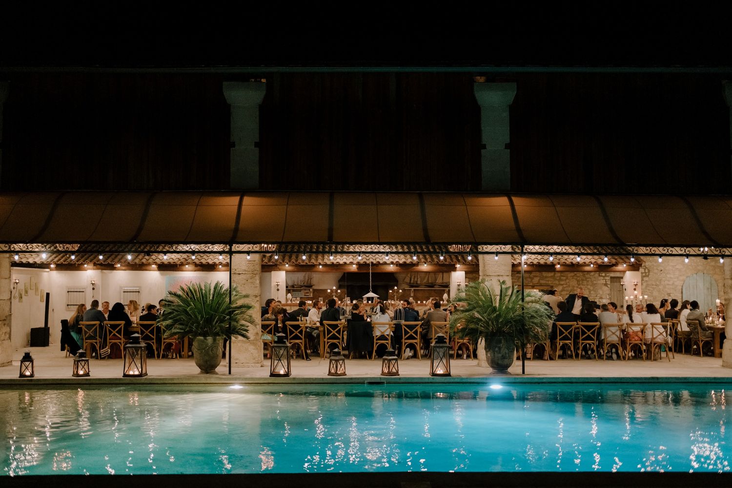 Nighttime view of an illuminated pool area with dining tables and string lights.