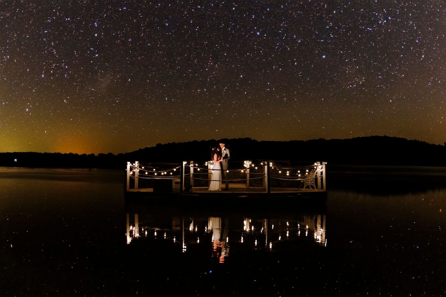 Wedding couple silhouetted against starry night sky with reflections on still water.